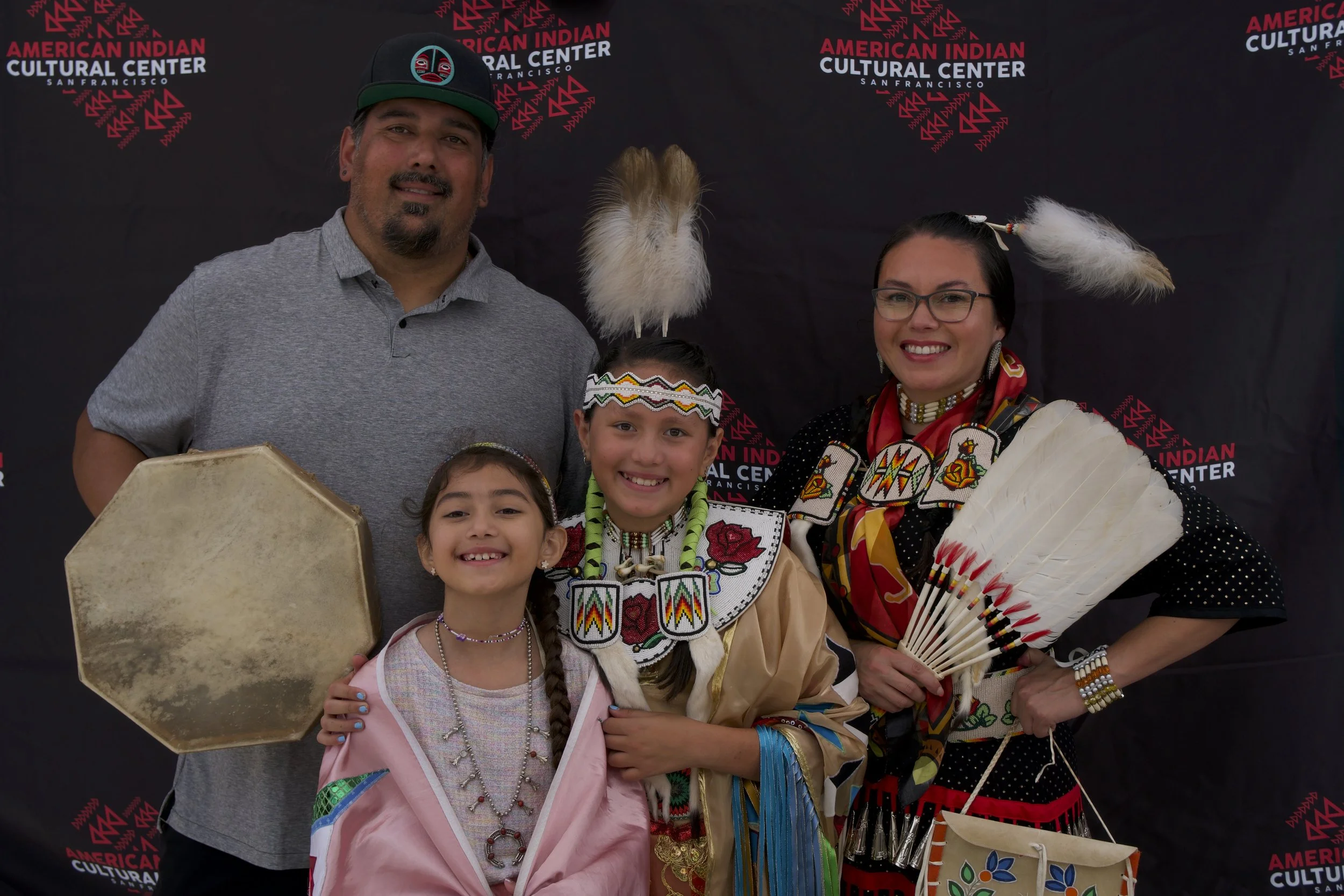 Four people, two children and two adults, dressed in traditional Native American attire, standing in front of a black backdrop with the words 'American Indian Cultural Center San Francisco' repeated in red and white text. One adult is holding a drum, and the woman on the right is holding a feather fan.
