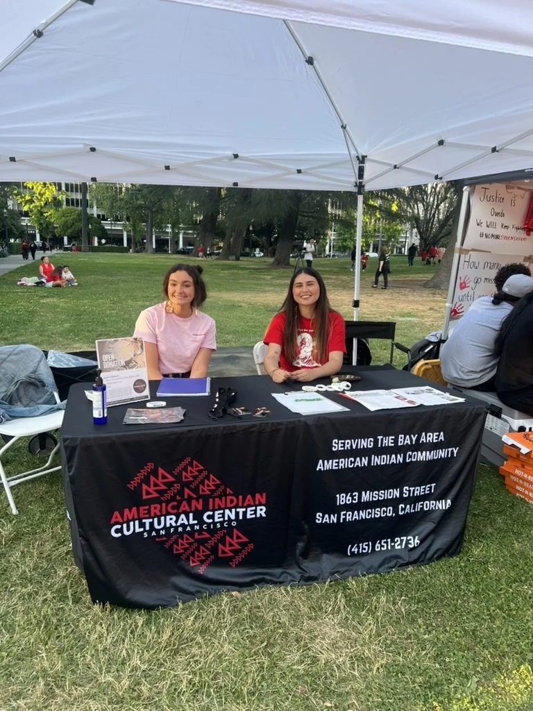 Two young women sitting behind a booth at an outdoor event, representing the American Indian Cultural Center in San Francisco, with a banner displaying their contact information and location.