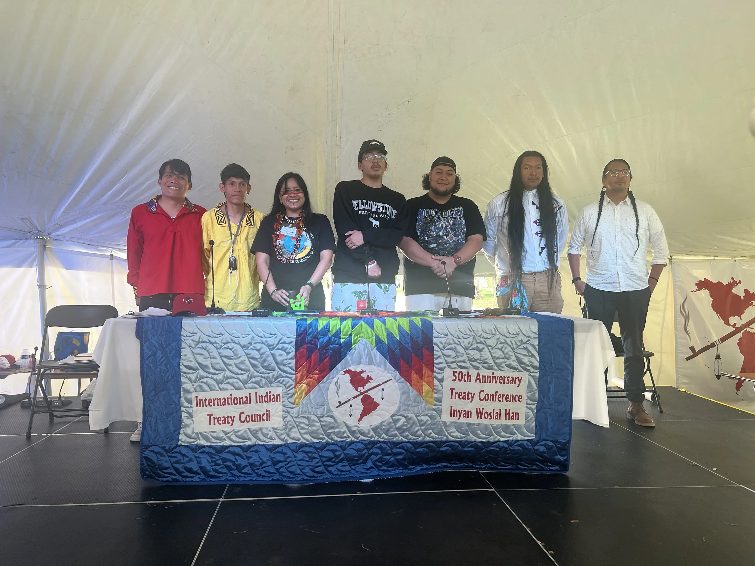 Seven people standing behind a table with a banner at an international treaty conference, inside a large tent. The banner reads 'International Indian Treaty Council' and '50th Anniversary Treaty Conference Iyan Wosla Han'. The group includes individuals of diverse backgrounds, dressed in casual and traditional attire.
