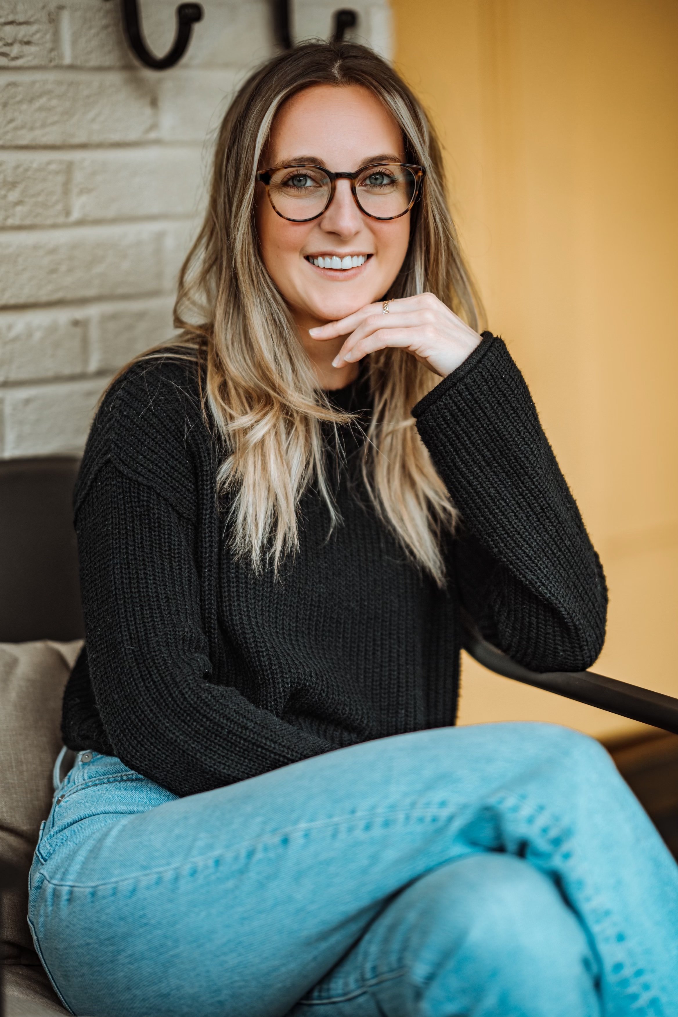 A young woman with blonde hair, glasses, wearing a black sweater and light blue jeans, smiling and sitting indoors.