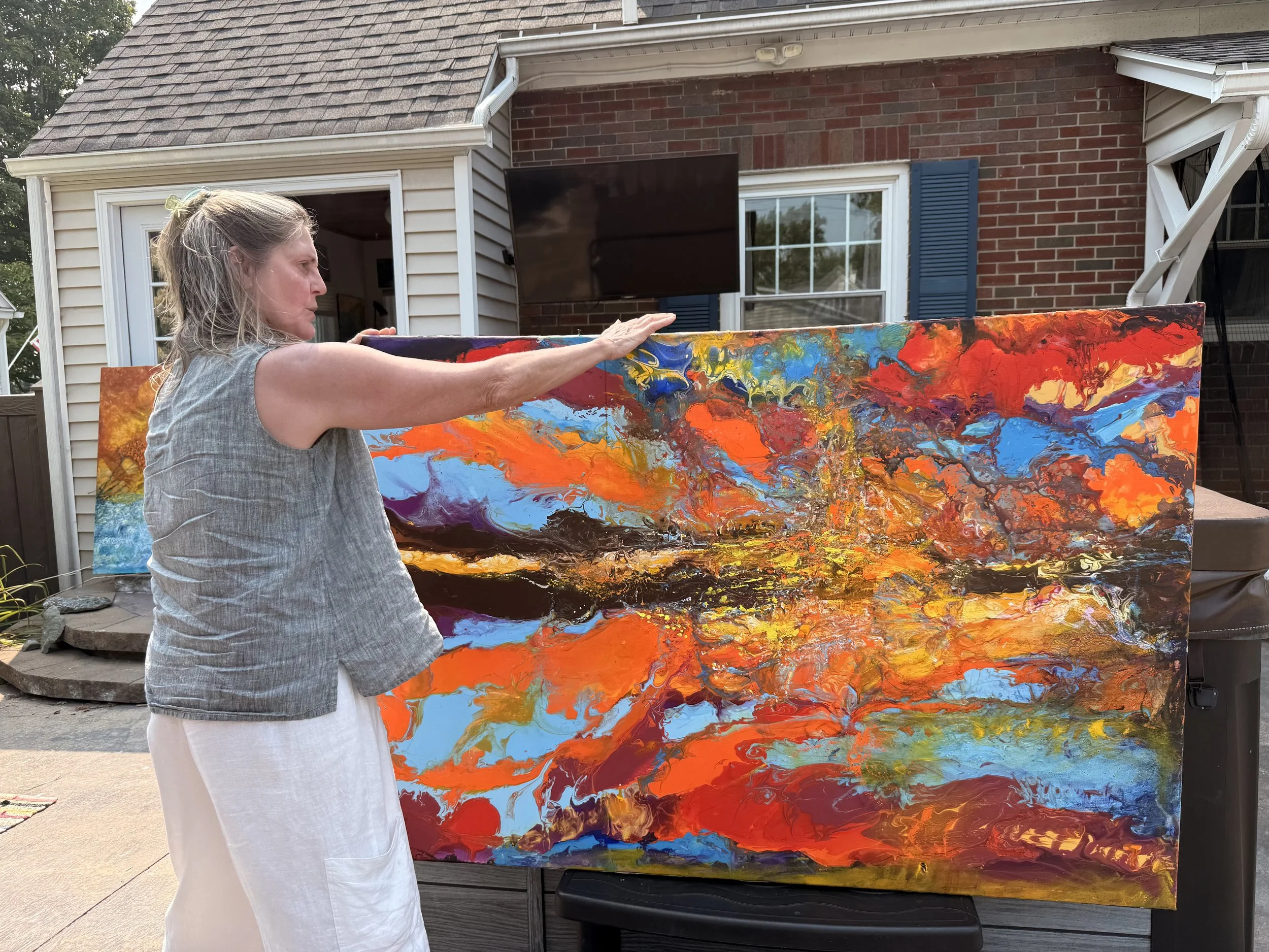 A woman is outdoors, standing in front of a large abstract painting with vibrant orange, red, yellow, black, blue, and purple colors. She is wearing a gray sleeveless top and white pants, and she appears to be exploring or adjusting the painting. There is a house with brick and siding exterior, a window, and a television in the background.
