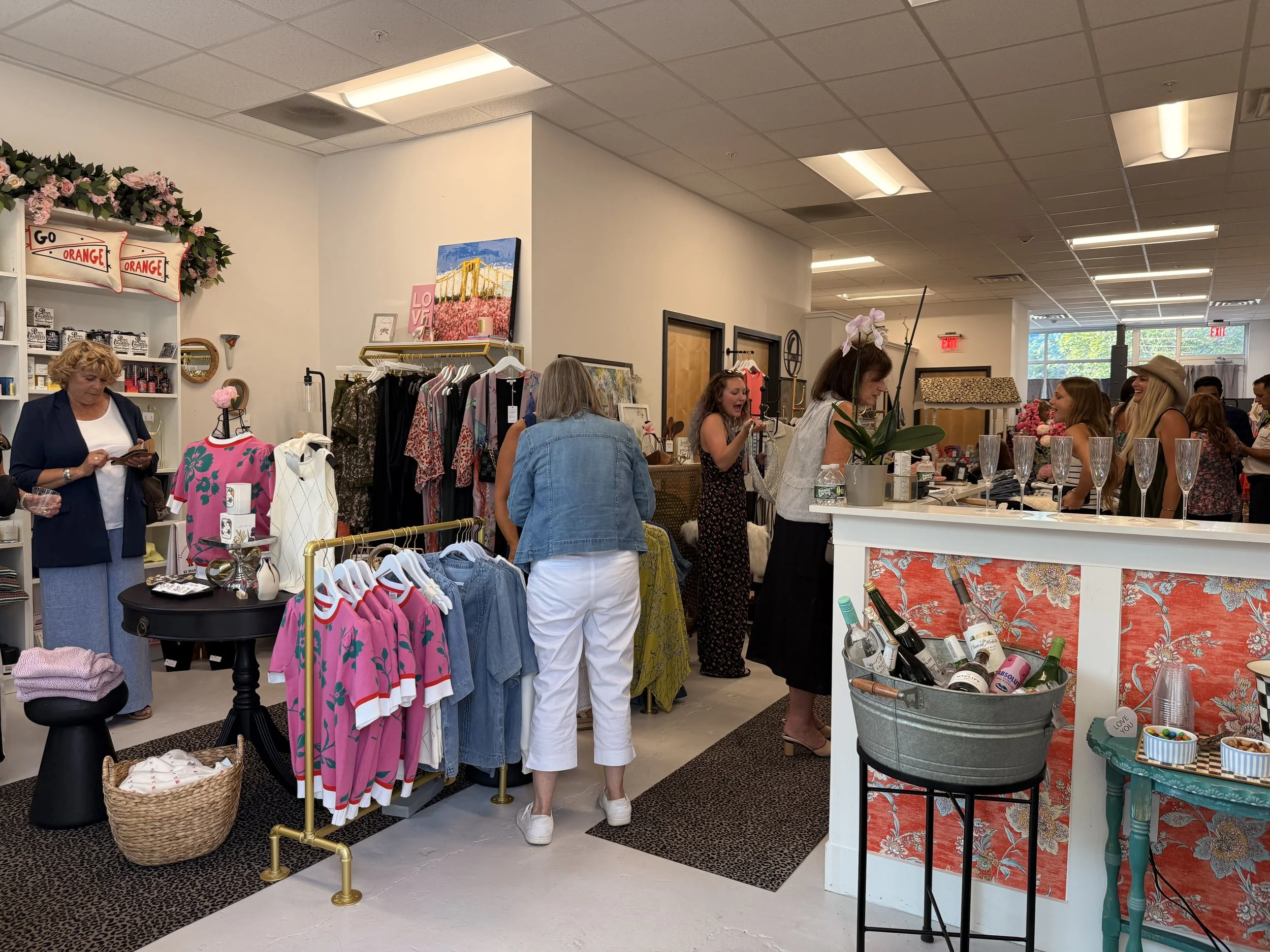 People shopping and socializing inside a boutique retail store with clothing, accessories, and drinks.