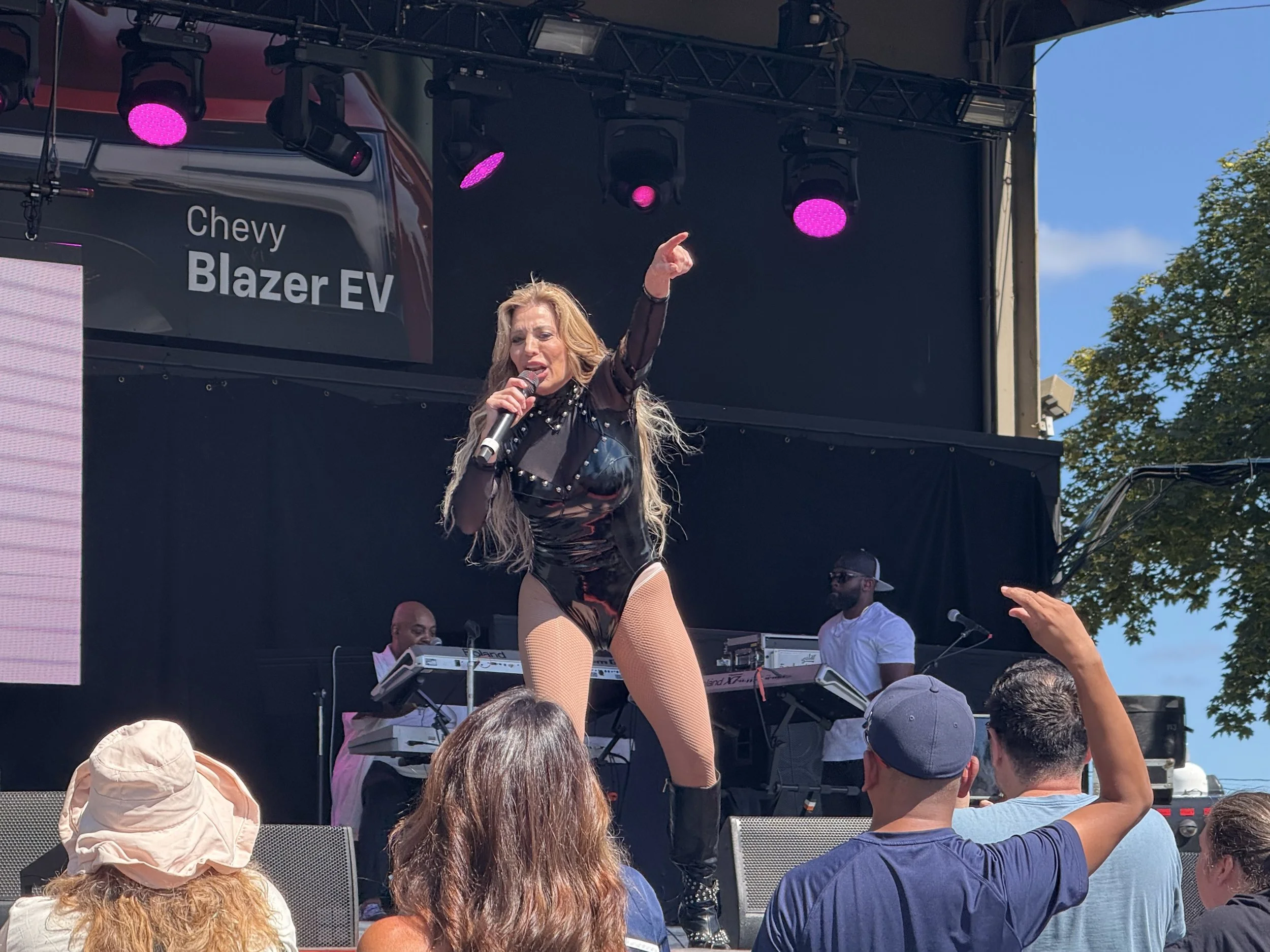 A female singer performing on an outdoor stage, holding a microphone with one arm raised, wearing a black leather outfit with fishnet stockings, black boots, and long wavy hair. There are two male musicians playing keyboards and guitar in the background. Audience members are watching, some raising their hands.