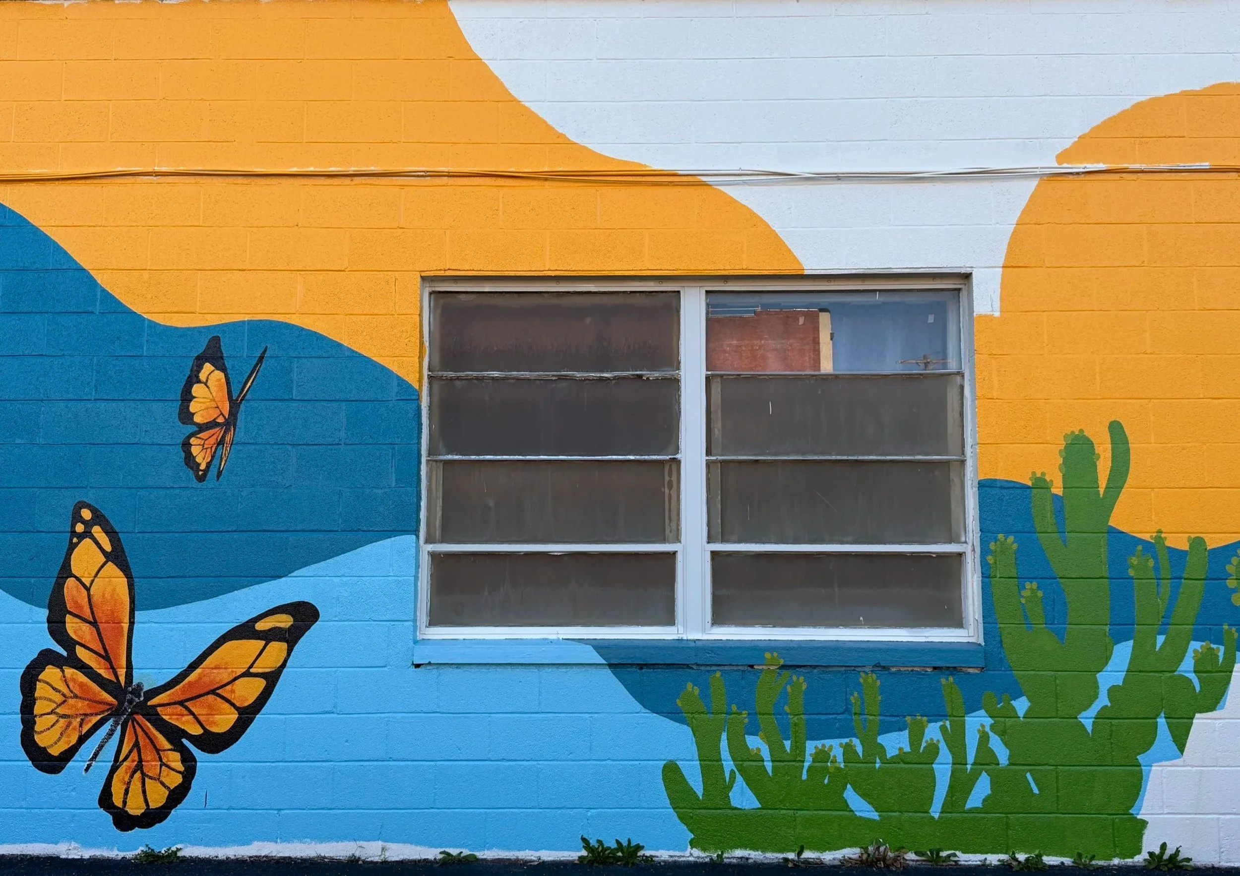 Mural painting on a brick wall with orange, blue, and white abstract shapes, green cactus, and orange butterflies next to a window.