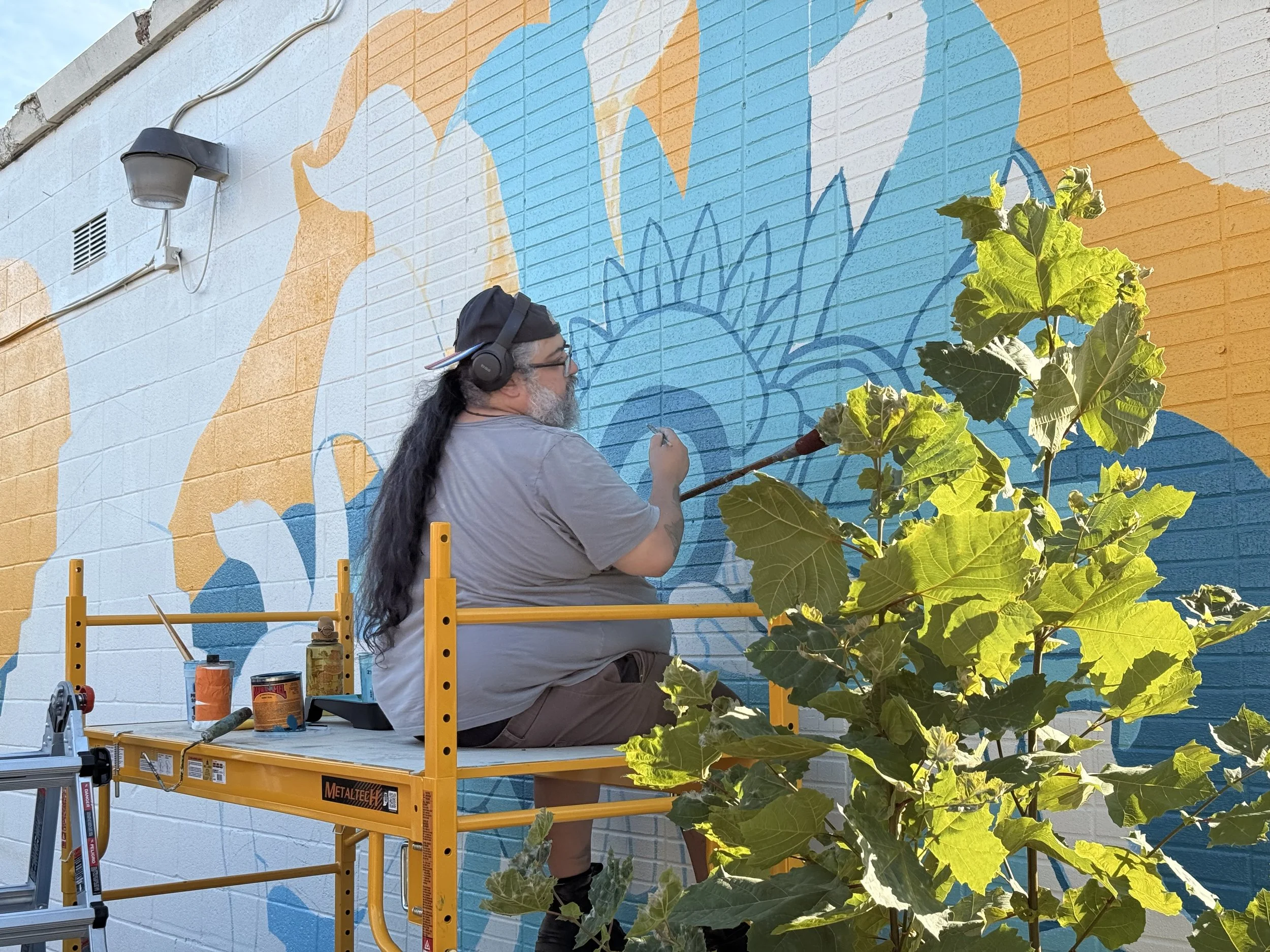 A man with long dark hair, glasses, and a beard painting a mural on a brick wall. He is sitting on a yellow scaffold, wearing headphones, a grey t-shirt, and a backward cap. The mural has large blue and orange shapes. A leafy green plant is in the foreground.