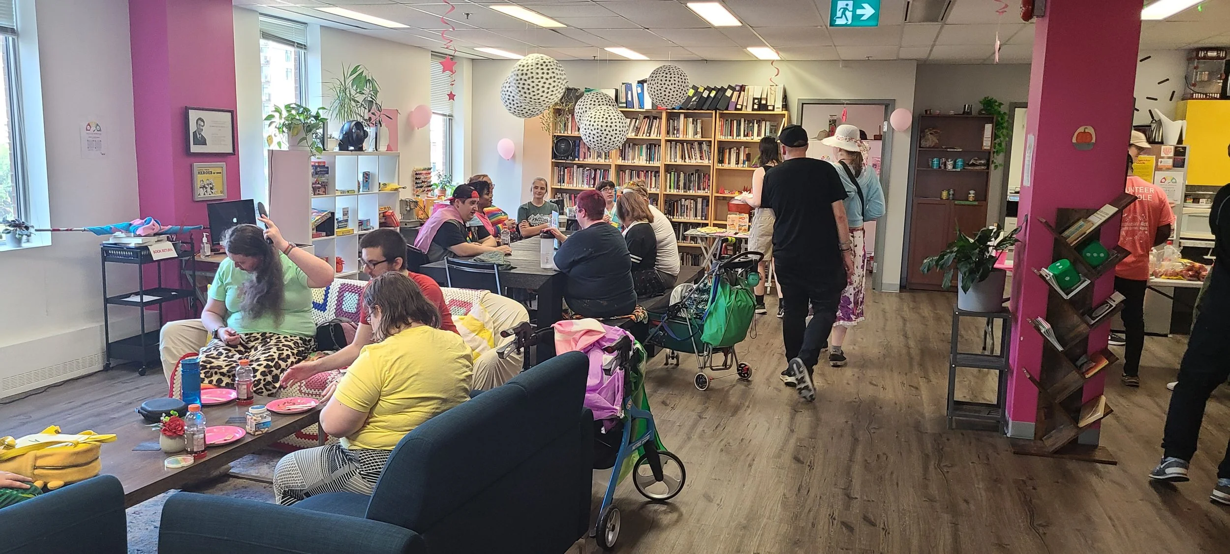 The interior of the Kind Space community centre, featuring a bright, open room with a large bookshelf filled with books. Several people are seated on comfortable sofas and chairs, engaged in conversation.