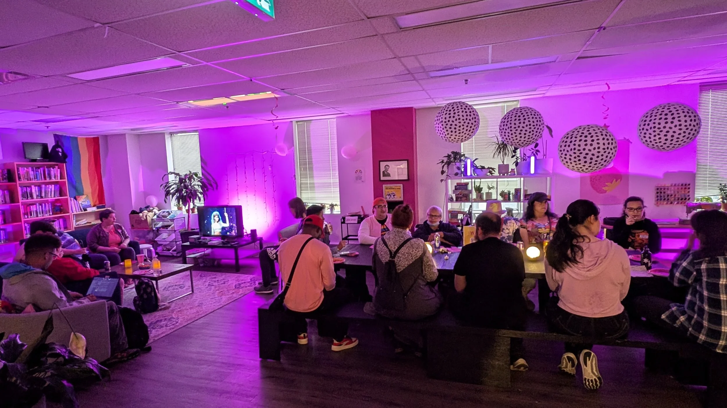 People gathered at a table in Kind Space under pink lights, pride flags, and patterned paper lanterns.