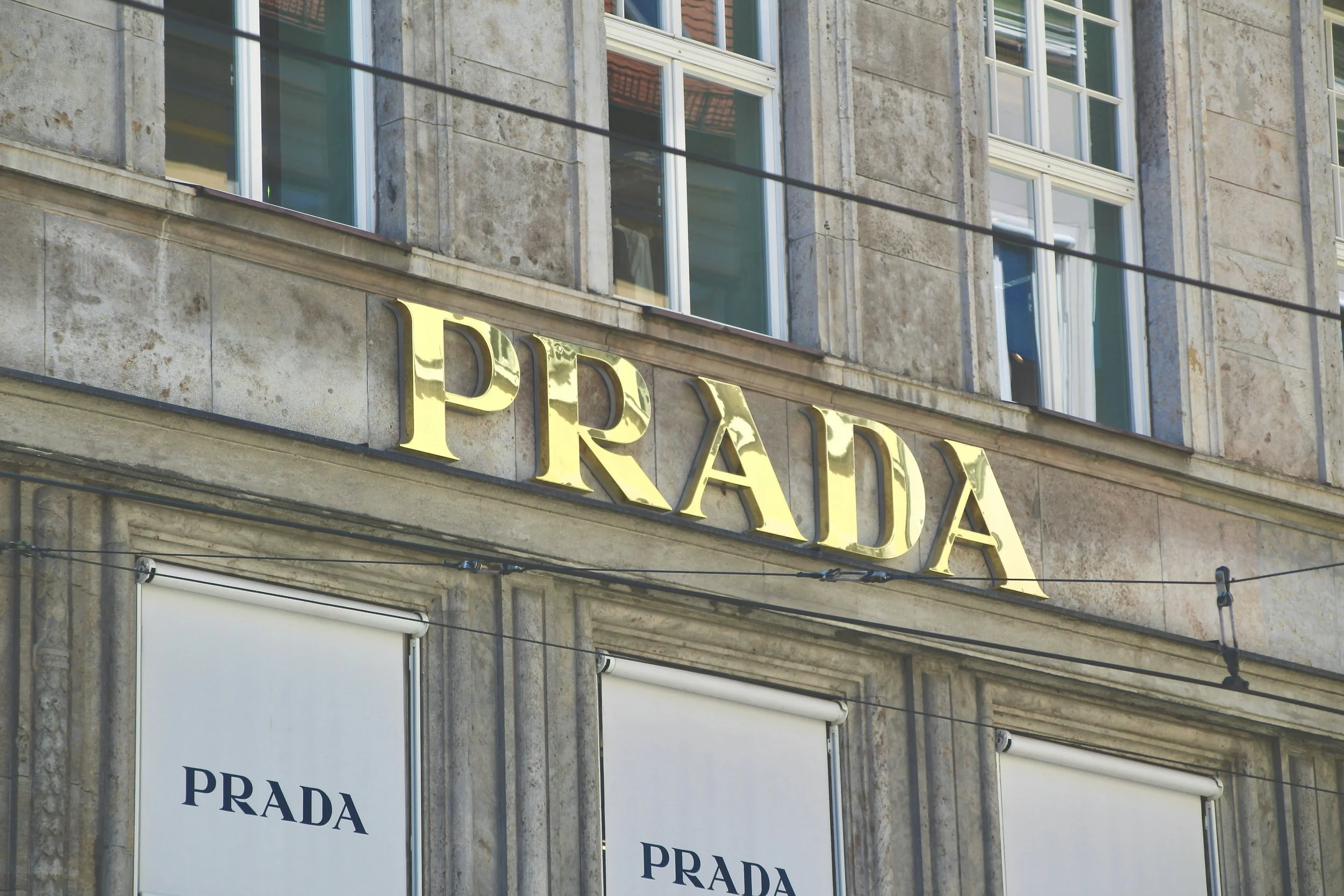 Exterior of a Prada store with gold-lettered Prada sign and fabric banners displaying Prada logo on large windows.