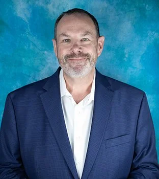 A man in a navy blue suit and white shirt smiling against a blue background.