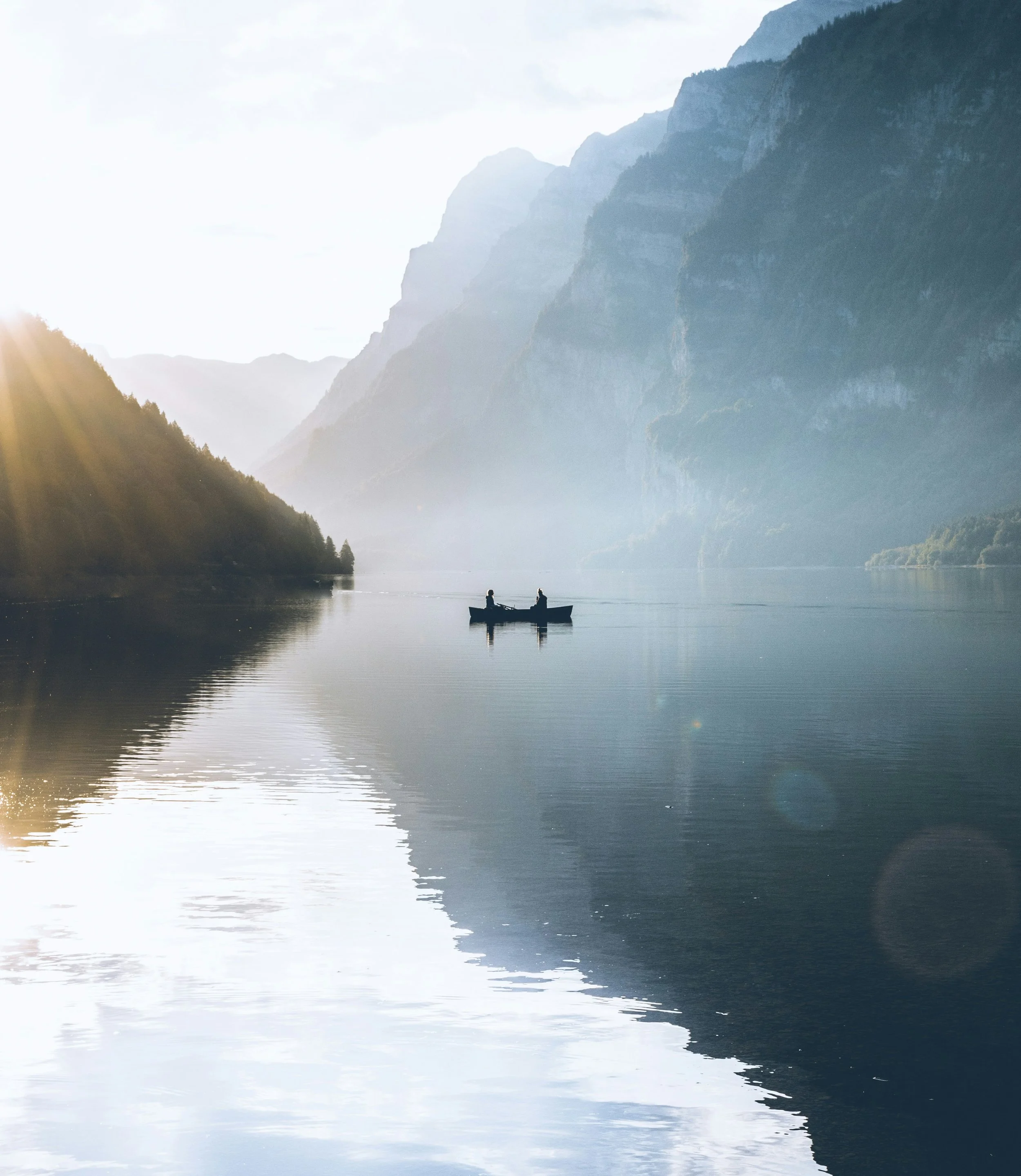 A peaceful lake surrounded by tall mountains with mist, and a boat with two people on the water, sunlight shining from the upper left.