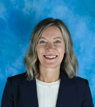 A woman with shoulder-length blonde hair, wearing a navy blazer and white top, smiling against a blue background.