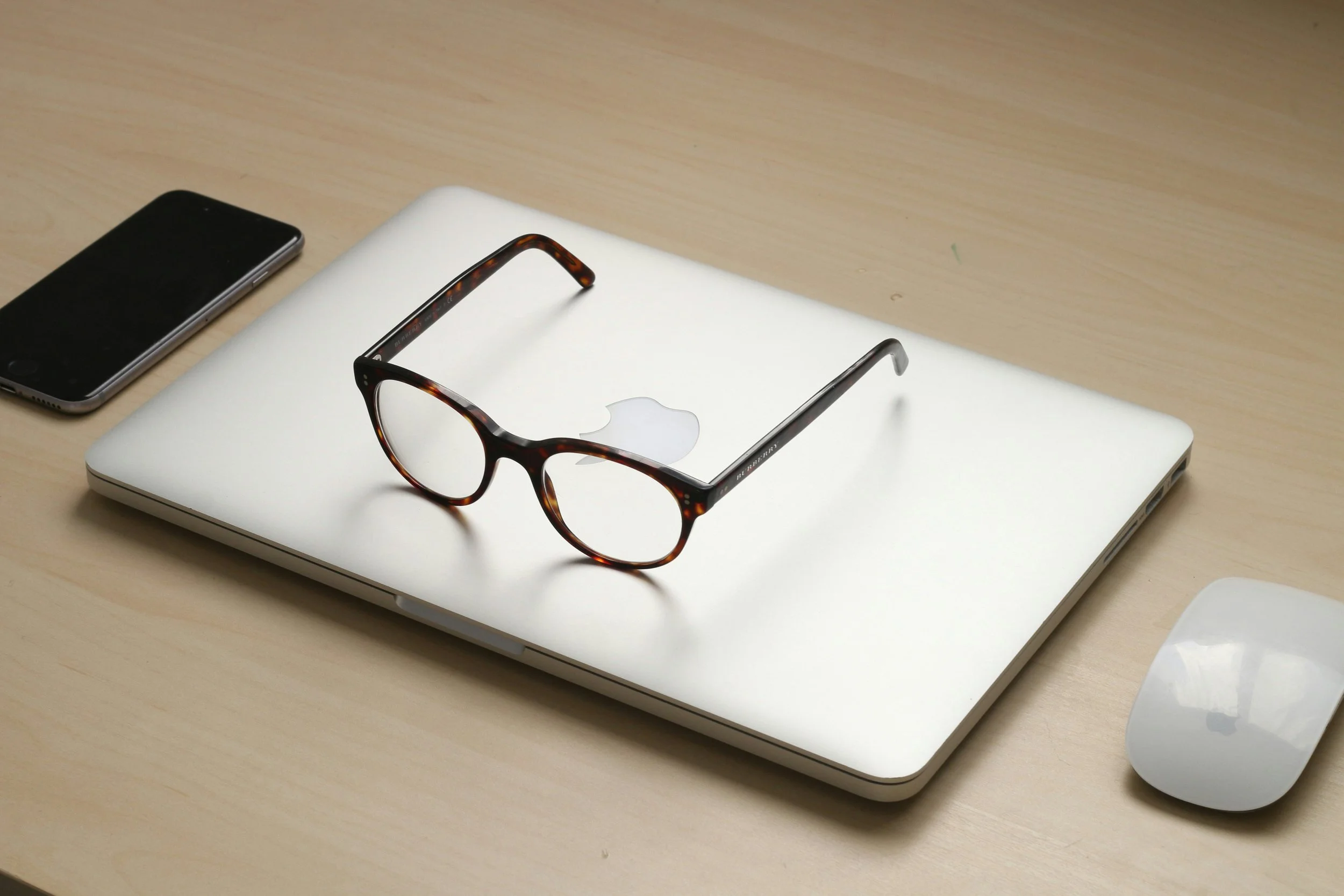 A closed silver MacBook with a pair of tortoiseshell glasses resting on top, a black smartphone to the left, and a white Apple Magic Mouse to the right, all on a light wood desk.
