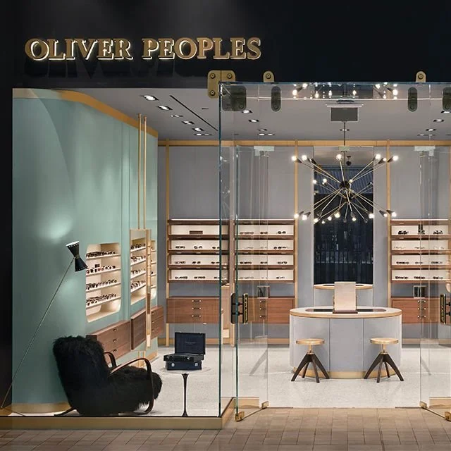 A modern eyewear store named Oliver Peoples, featuring isolation glass walls, wooden display shelves with sunglasses, a reception desk, and a stylish black chair in a cozy corner.