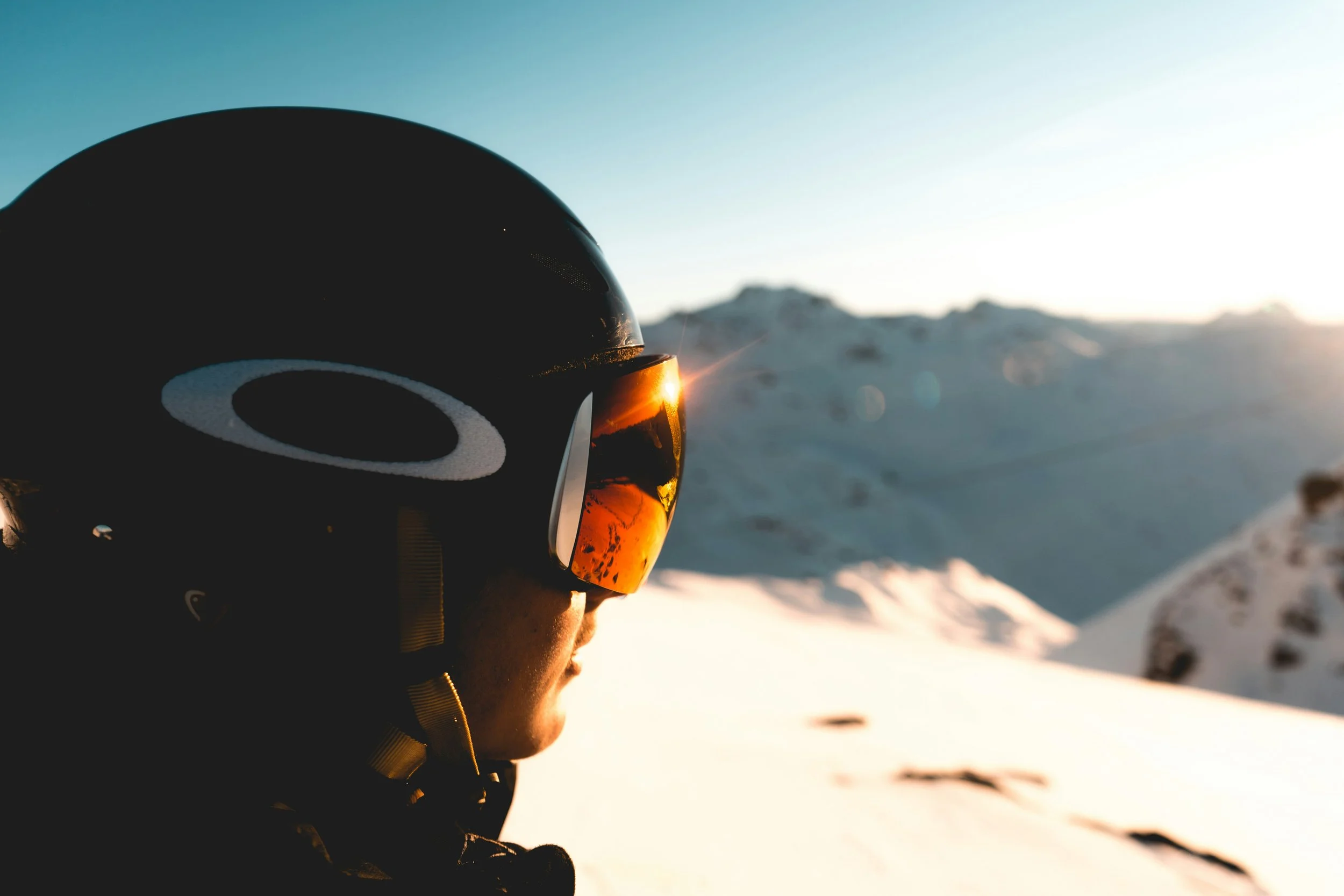 A person wearing a black helmet and ski goggles, staring at a snowy mountain landscape during sunset.