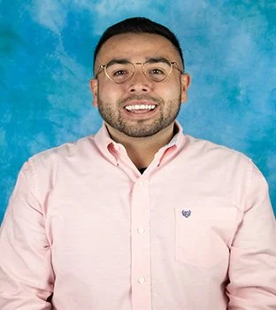 Portrait of a man with short dark hair and glasses, smiling, wearing a light pink button-up shirt, against a blue textured background.