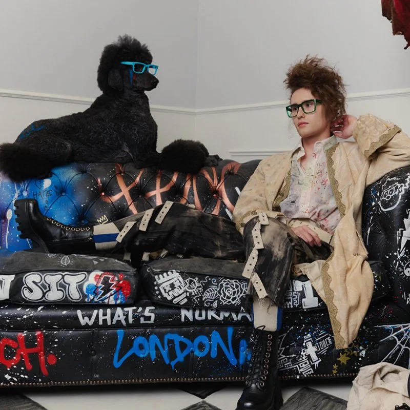 A person with curly hair wearing glasses, a patterned shirt, and a beige jacket, sitting on a graffiti-covered black couch, looking at a black poodle dog dressed in a jacket, with blue glasses and a matching blue bowtie, on the couch beside them.