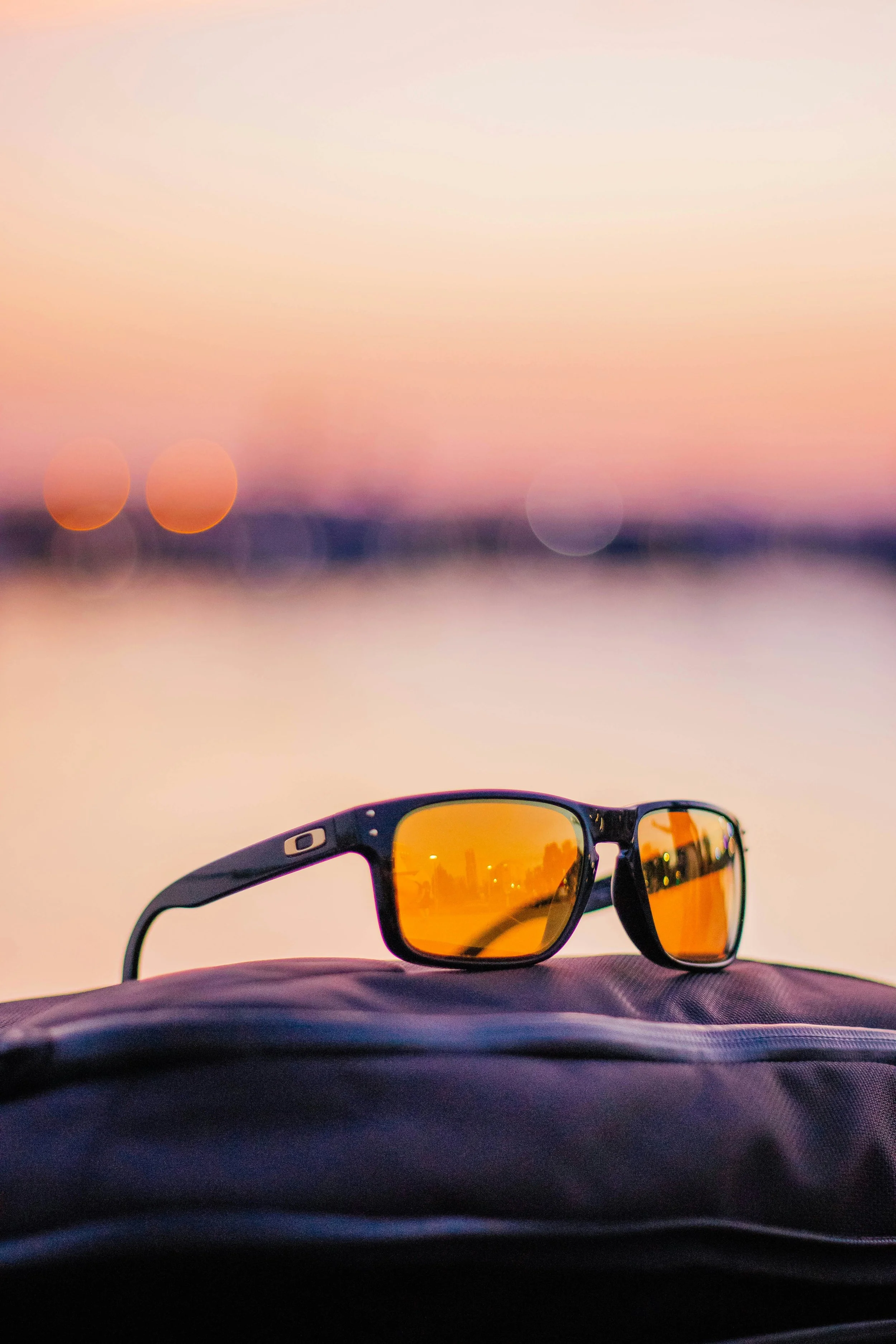 Sunglasses with orange lens reflecting a city skyline, resting on a fabric surface during sunset with blurred background of water and colorful sky.