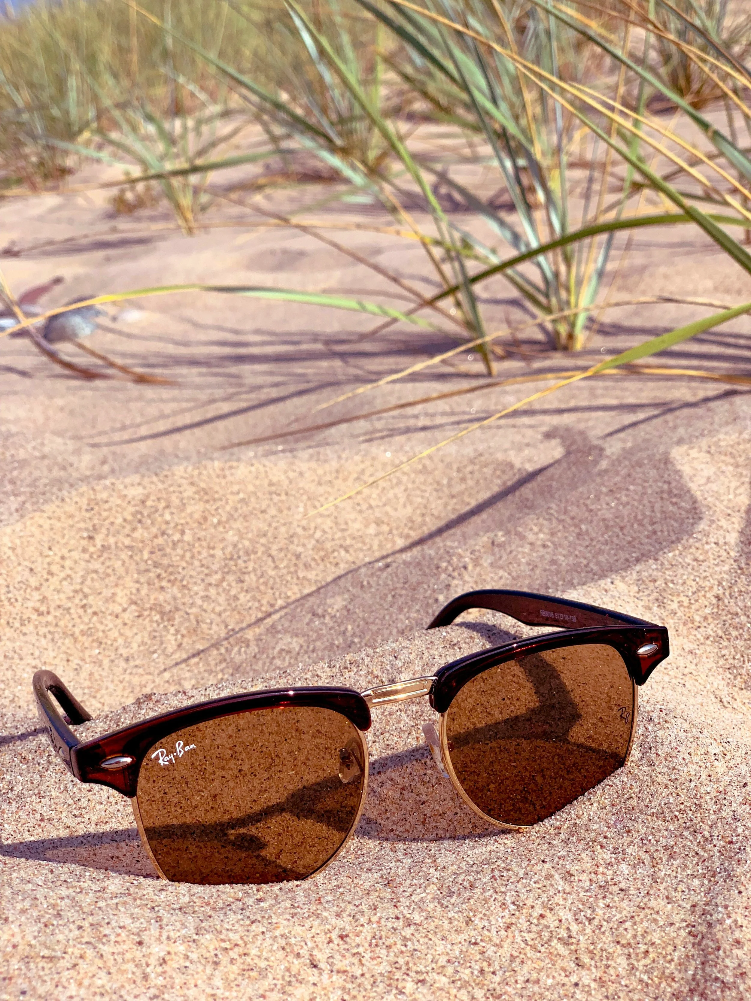 A pair of Ray-Ban sunglasses with dark lenses lying on sandy ground at the beach, with grass in the background.