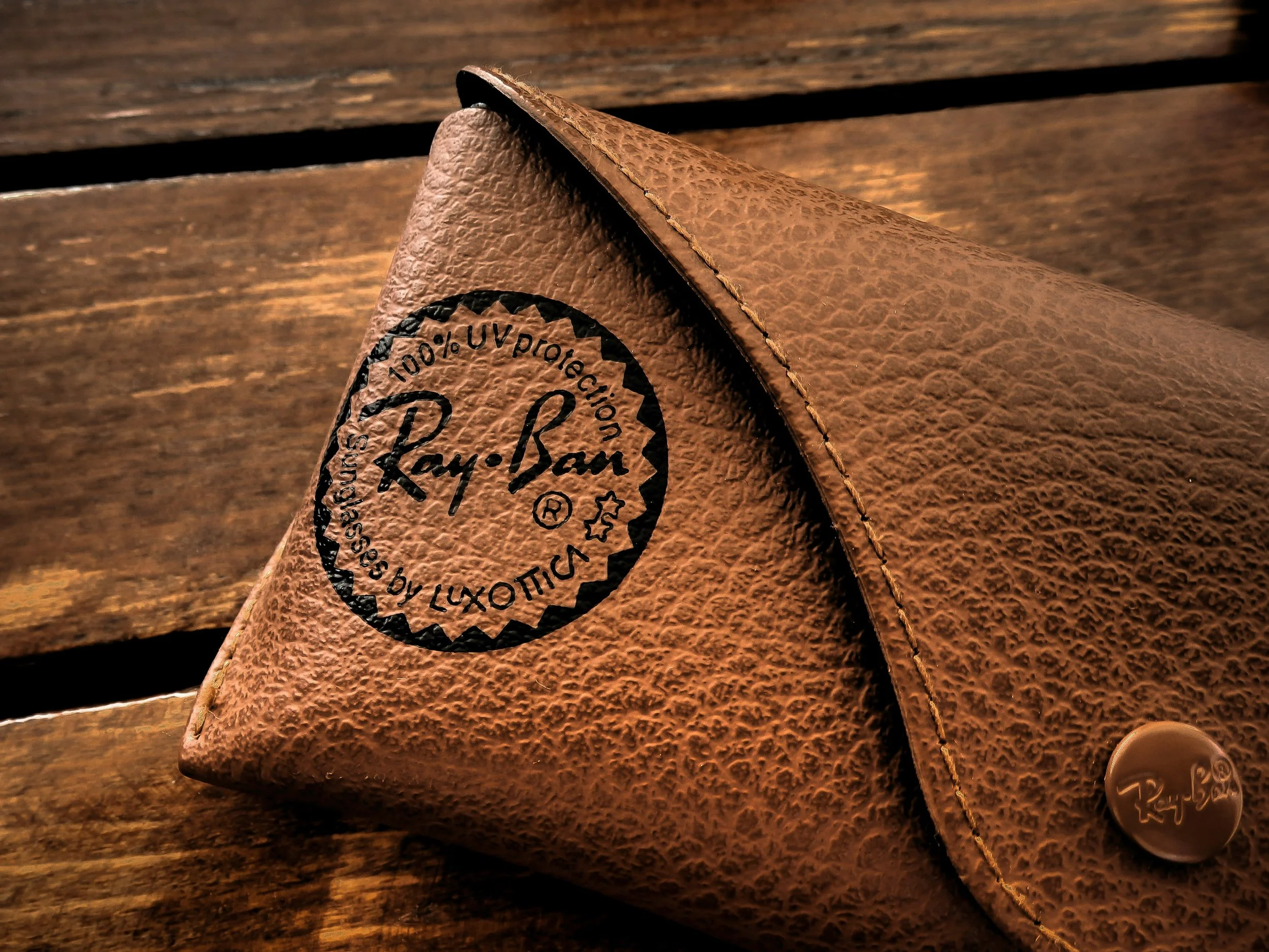 Brown leather Ray-Ban sunglasses case with logo and authenticity stamp on a wooden surface.
