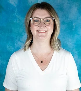 Portrait of a smiling woman with blonde hair, wearing glasses and a white top, against a blue background.