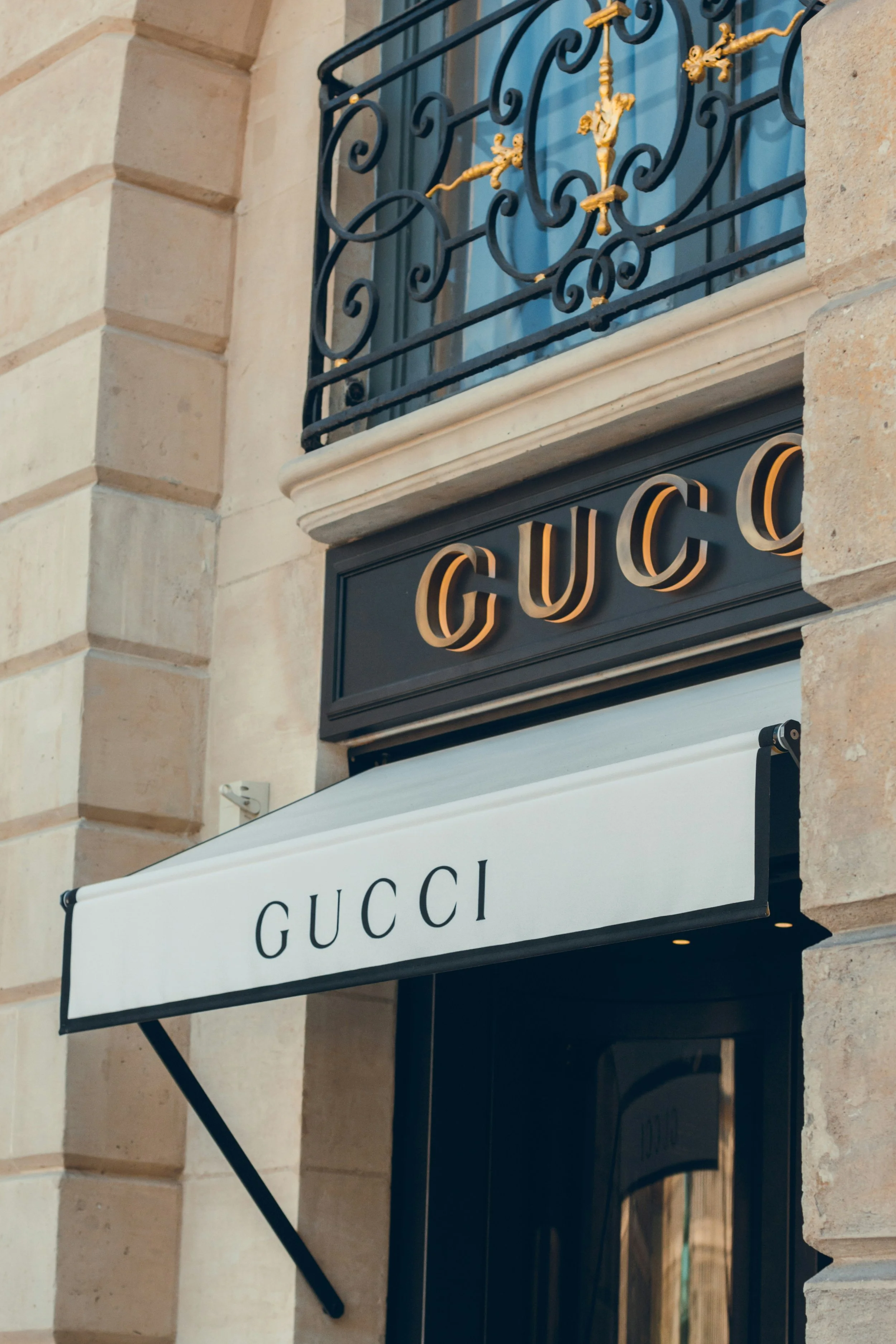 Storefront of Gucci with a black sign, gold lettering, and a white awning with black text, on a beige stone building.