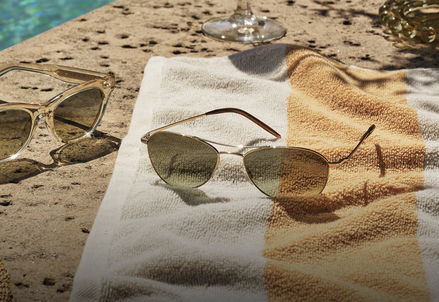 A pair of sunglasses resting on a striped towel on a sandy surface near a pool.