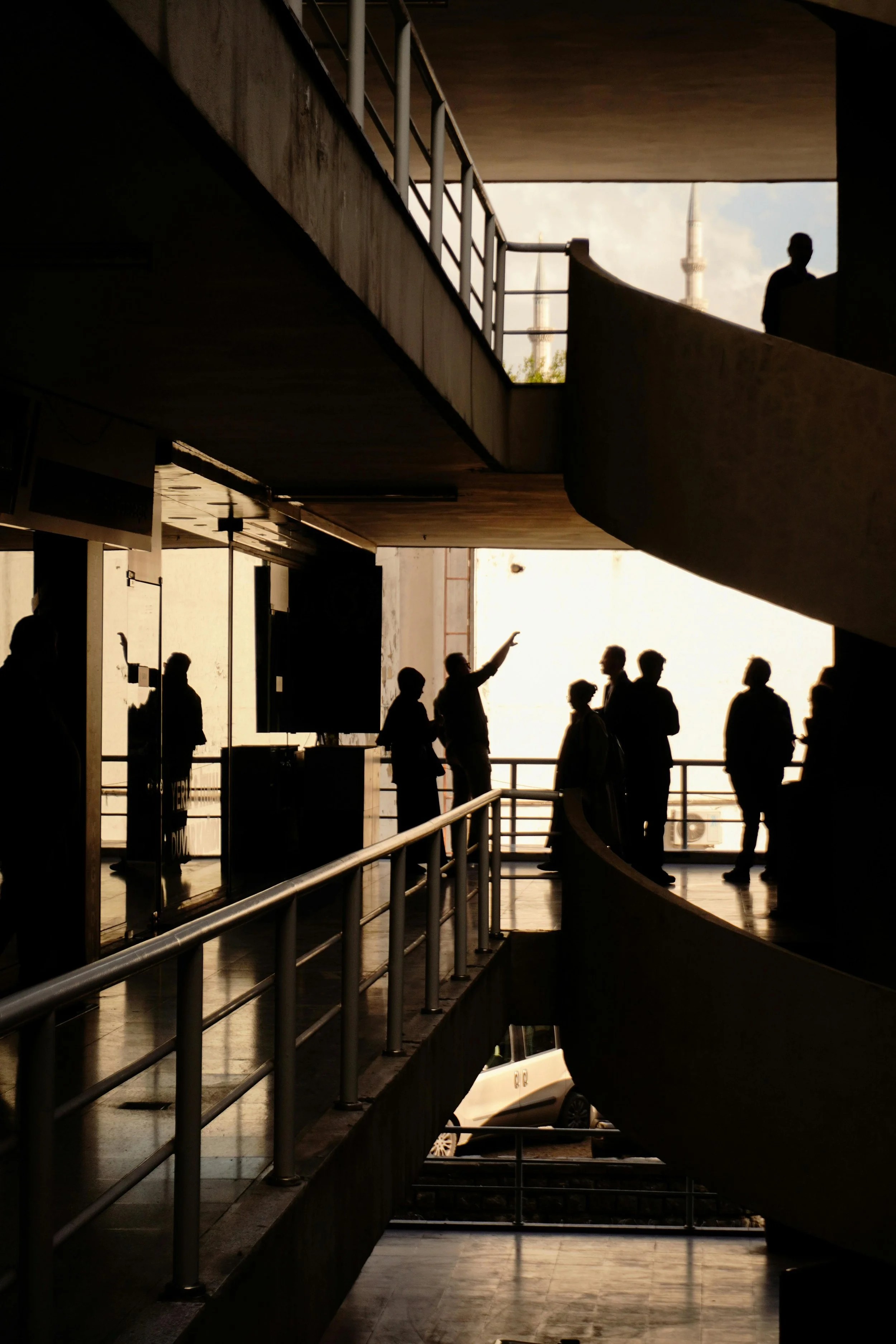 Silhouettes of people on an indoor balcony overlooking an open area, with a person gesturing and others in conversation, against a bright outdoor background visible through large windows.