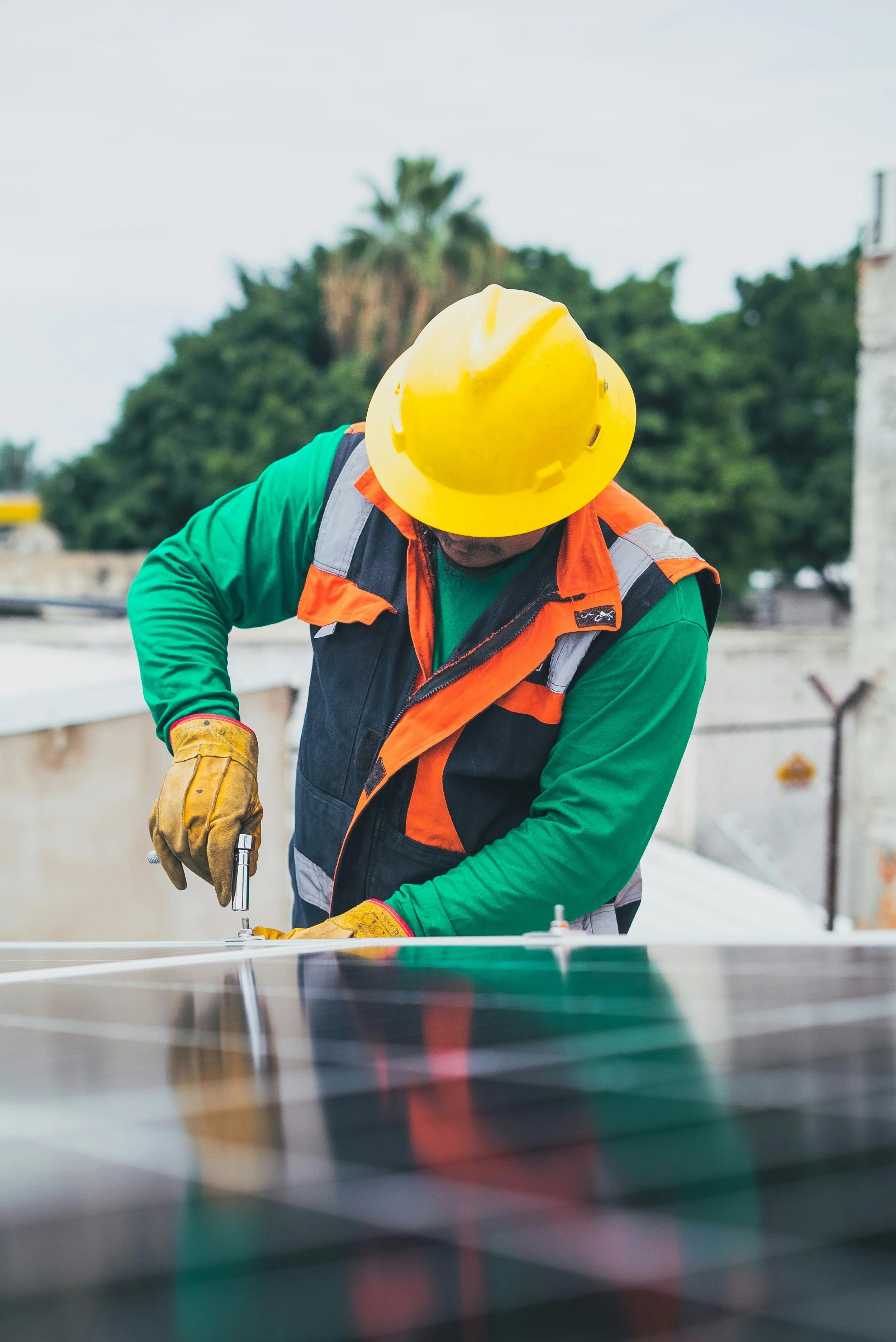 Worker installing or repairing solar panels on a rooftop, wearing a yellow safety helmet, orange and black vest, green long sleeve shirt, and yellow gloves.