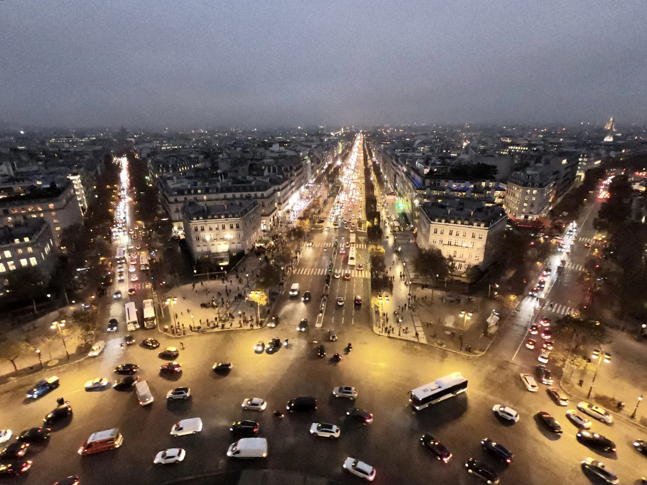 Aerial view of a city at dusk, showing busy streets with moving cars, buildings with lit windows, and a line of lights down the center of the city.