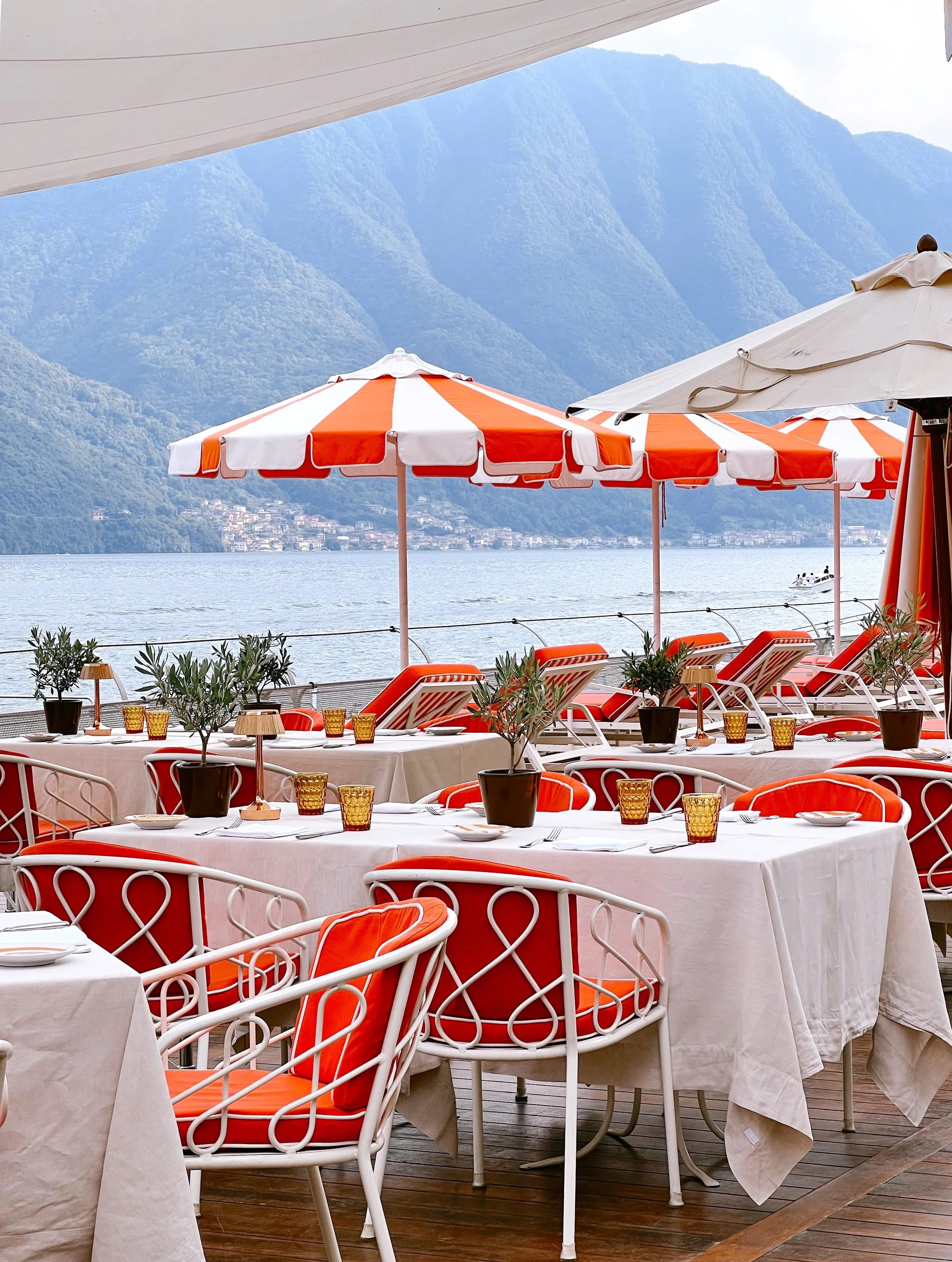 Outdoor restaurant with white tablecloths, orange chairs, potted plants, and orange umbrellas overlooking a body of water with mountains in the background. Amalfi Coast Lake Como Italy Columbus, Mississippi Travel Advisor Travel Agent Starkville, MS