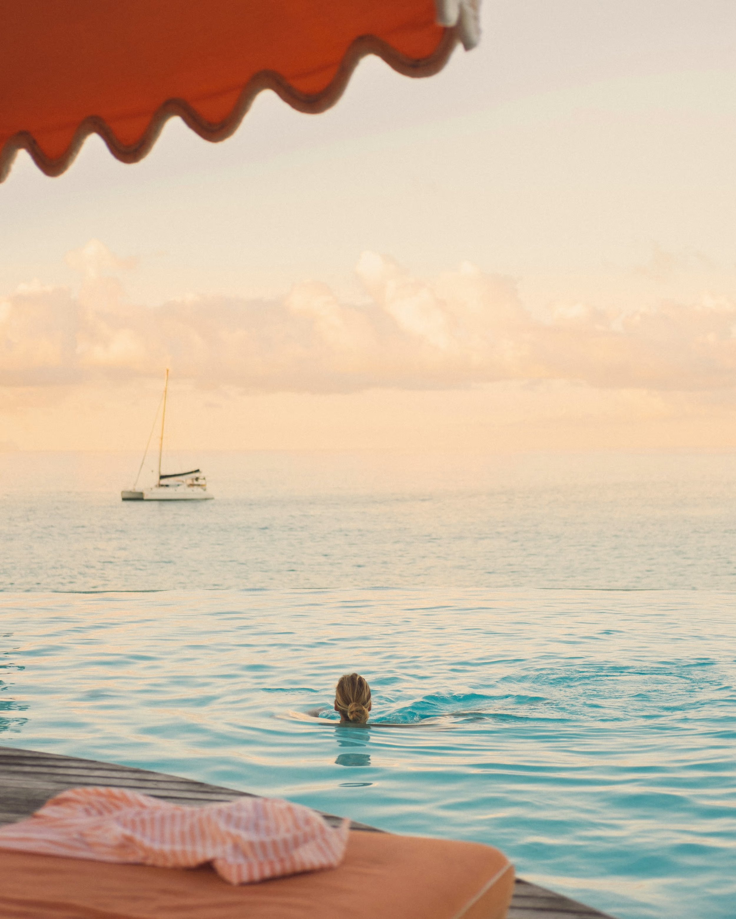 A person swimming in the ocean near a dock, with a boat in the distance and clouds in the sky, viewed from under a striped red and white awning. Luxury Belmond.