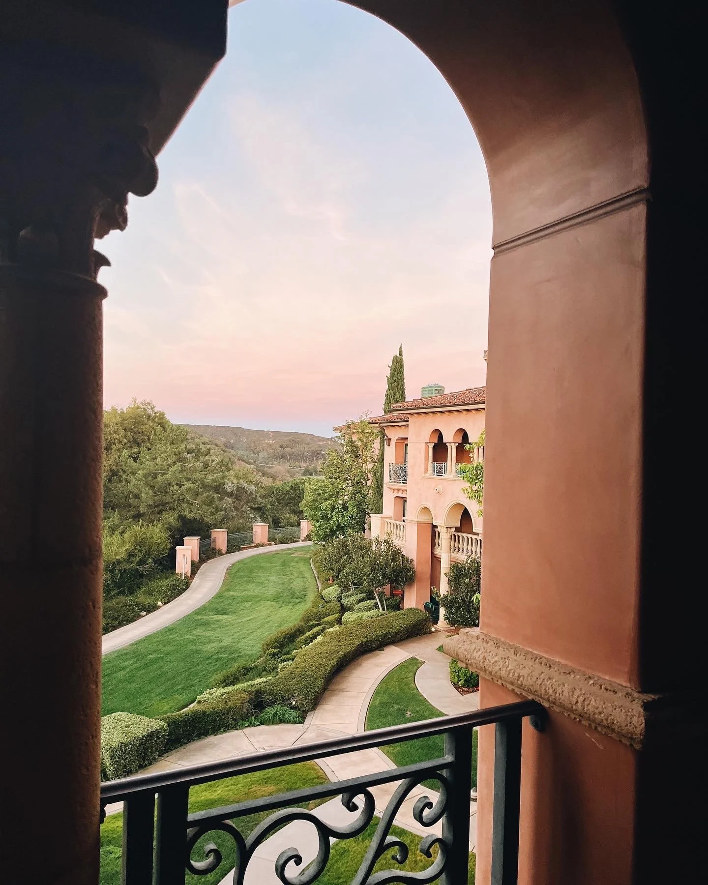 View of a lush green landscape with winding path, trees, and a pink building with balconies, seen from a balcony with decorative ironwork, at sunset