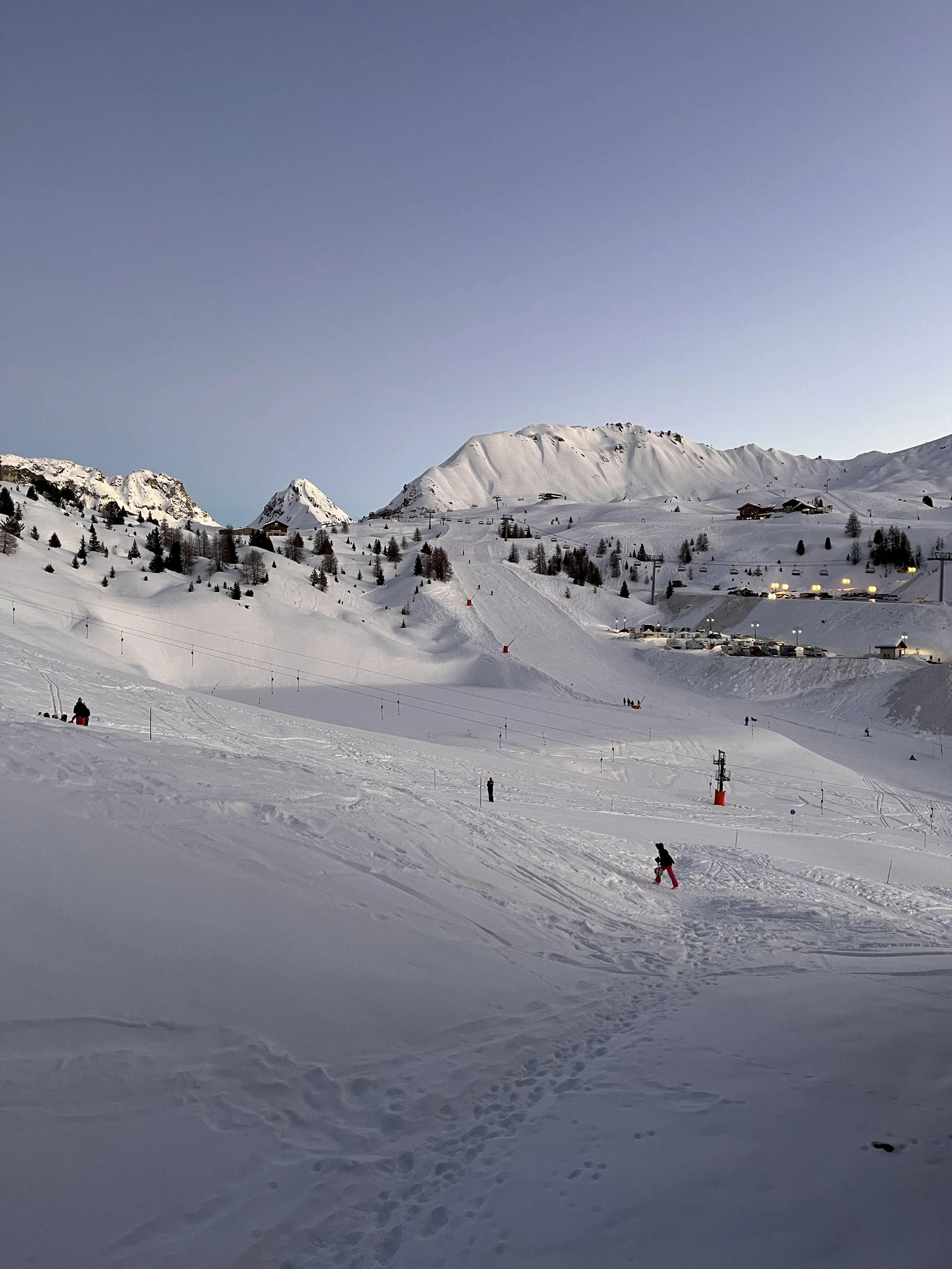 Snow-covered ski slopes with few skiers, ski lifts, small buildings, and mountains in the background during dusk.