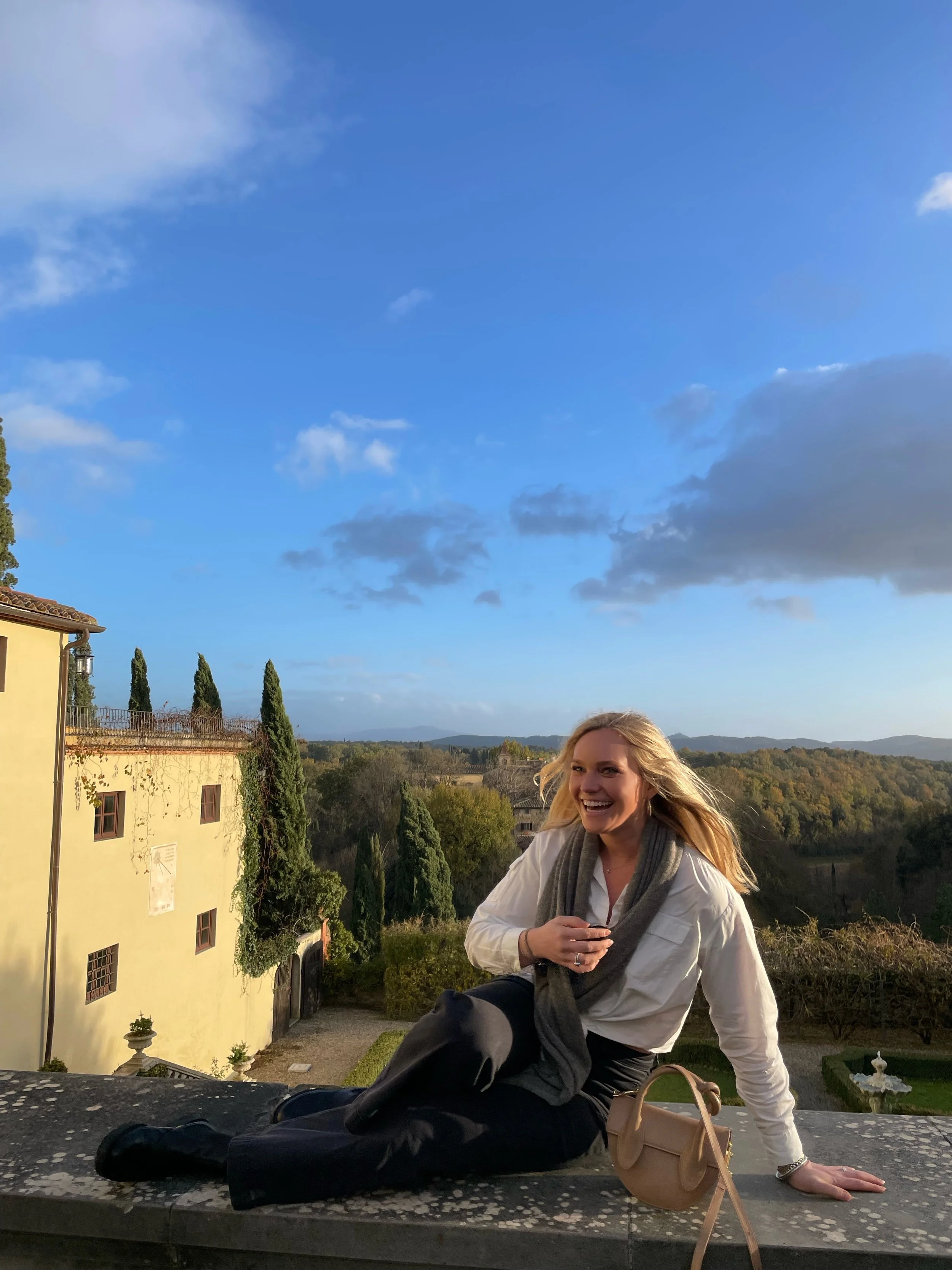 A woman with blonde hair sitting on a wall outdoors, smiling, with a scenic background of trees, hills, and a blue sky with clouds.