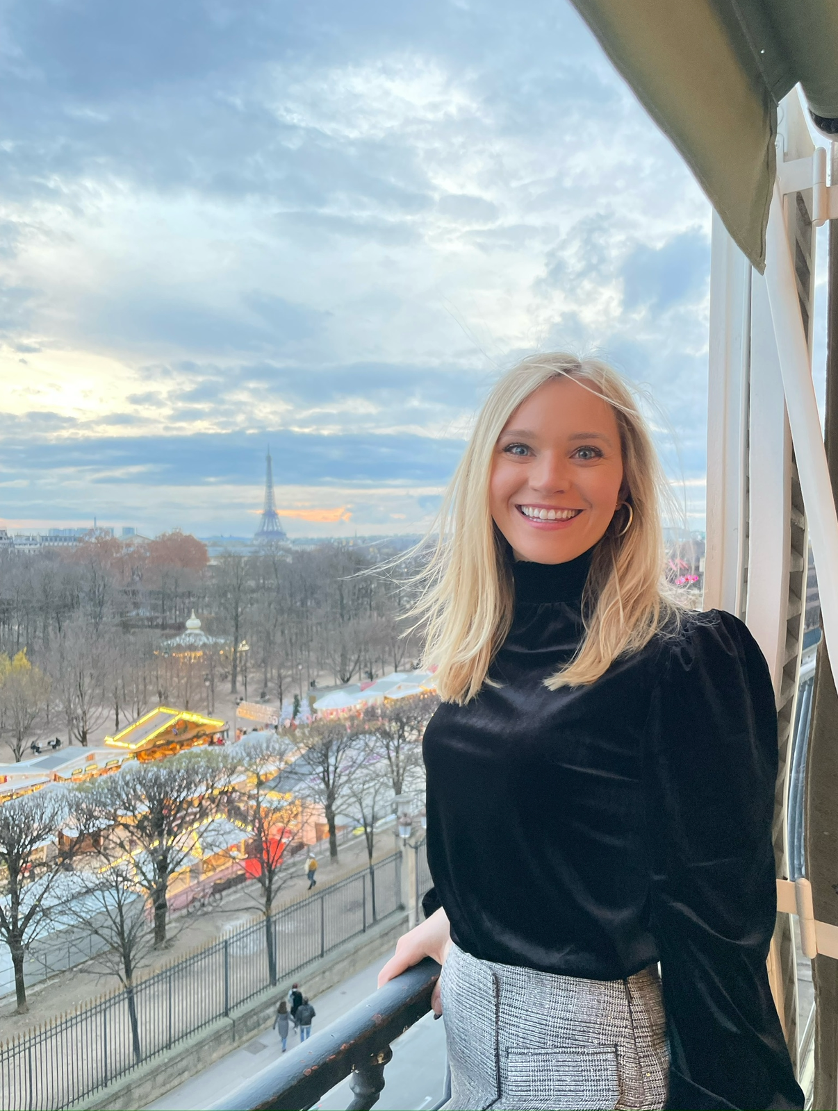 Smiling woman with blonde hair wearing a black top and plaid skirt standing on a balcony with a view of a park, trees, the Eiffel Tower, and a cloudy sky in Paris.