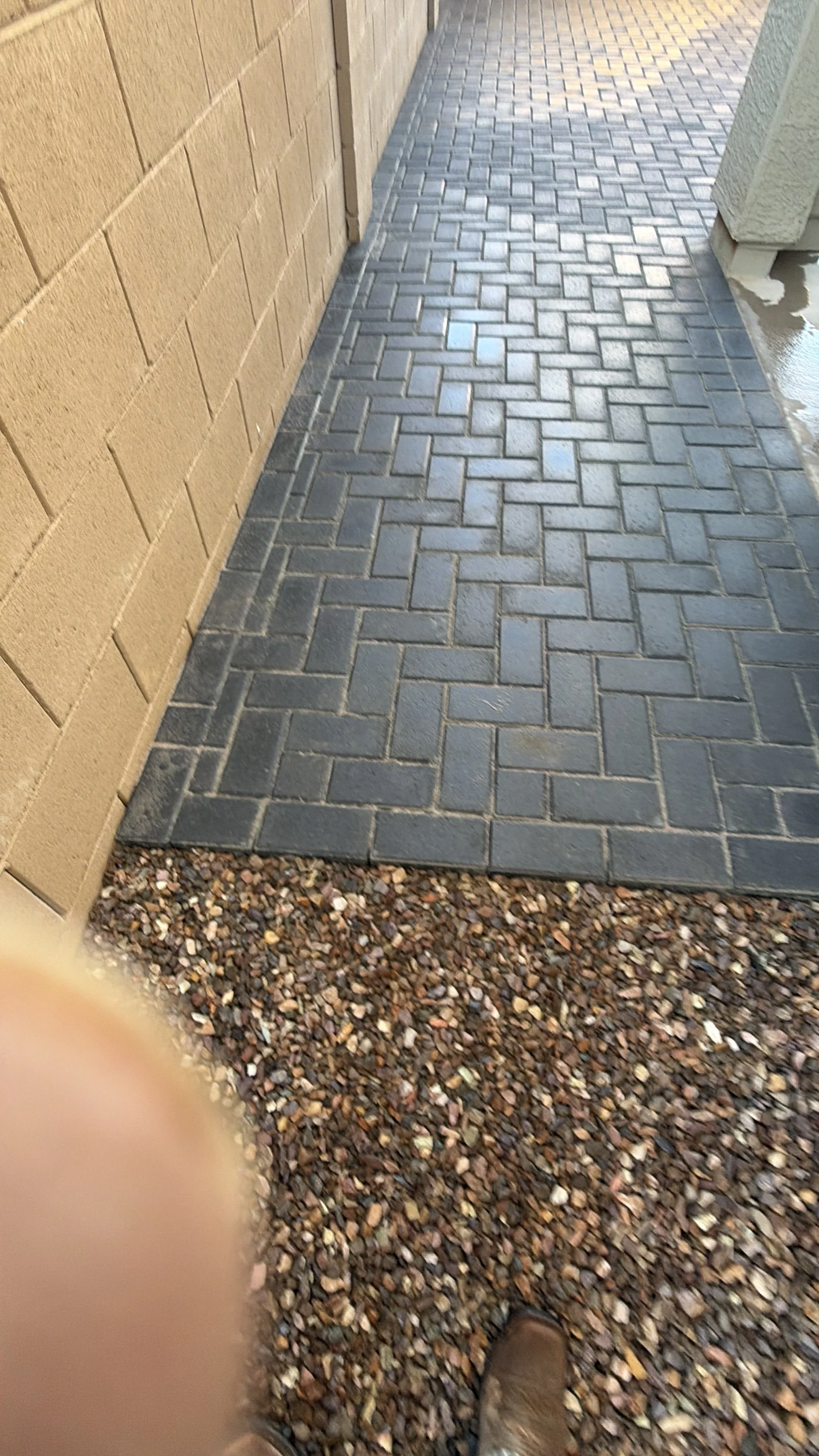 Photograph of a paved walkway with gray bricks laid in a herringbone pattern, bordered by a gravel area and beige brick walls on each side.