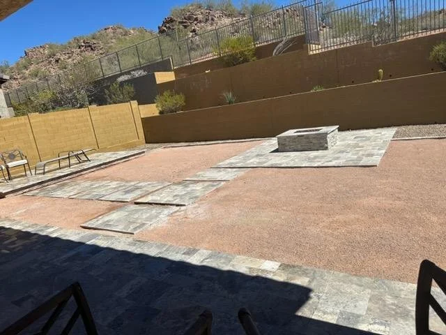 Outdoor backyard area with a paved pathway, benches, and a fire pit on a raised stone platform, surrounded by a brown wooden retaining wall and a chain-link fence at the top, with rocky hills in the background under a blue sky.