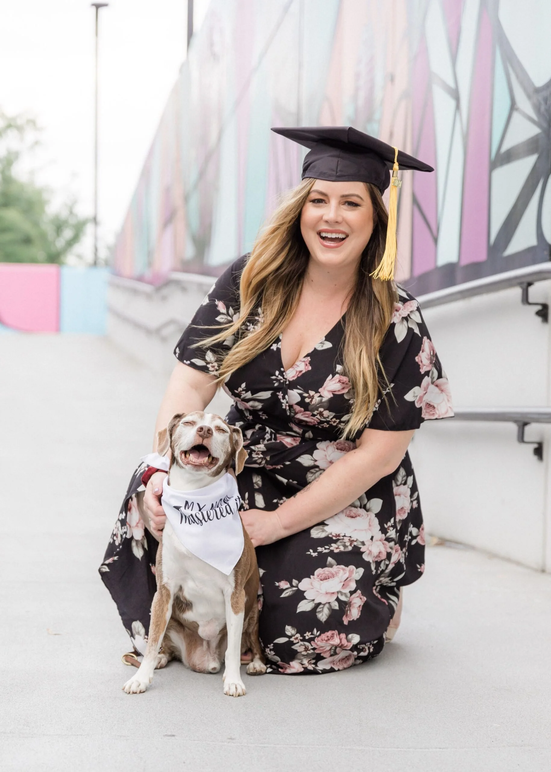 A woman in a floral dress and graduation cap kneeling beside a happy dog with a white bandana that says 'Mastered.' The woman is smiling, and the background features a colorful mural.