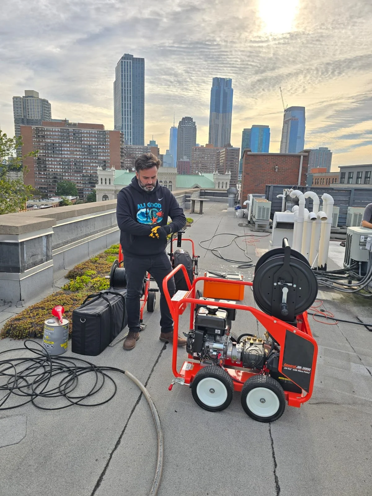 Man in blue uniform and gloves operating orange drain cleaning machine against beige brick wall.