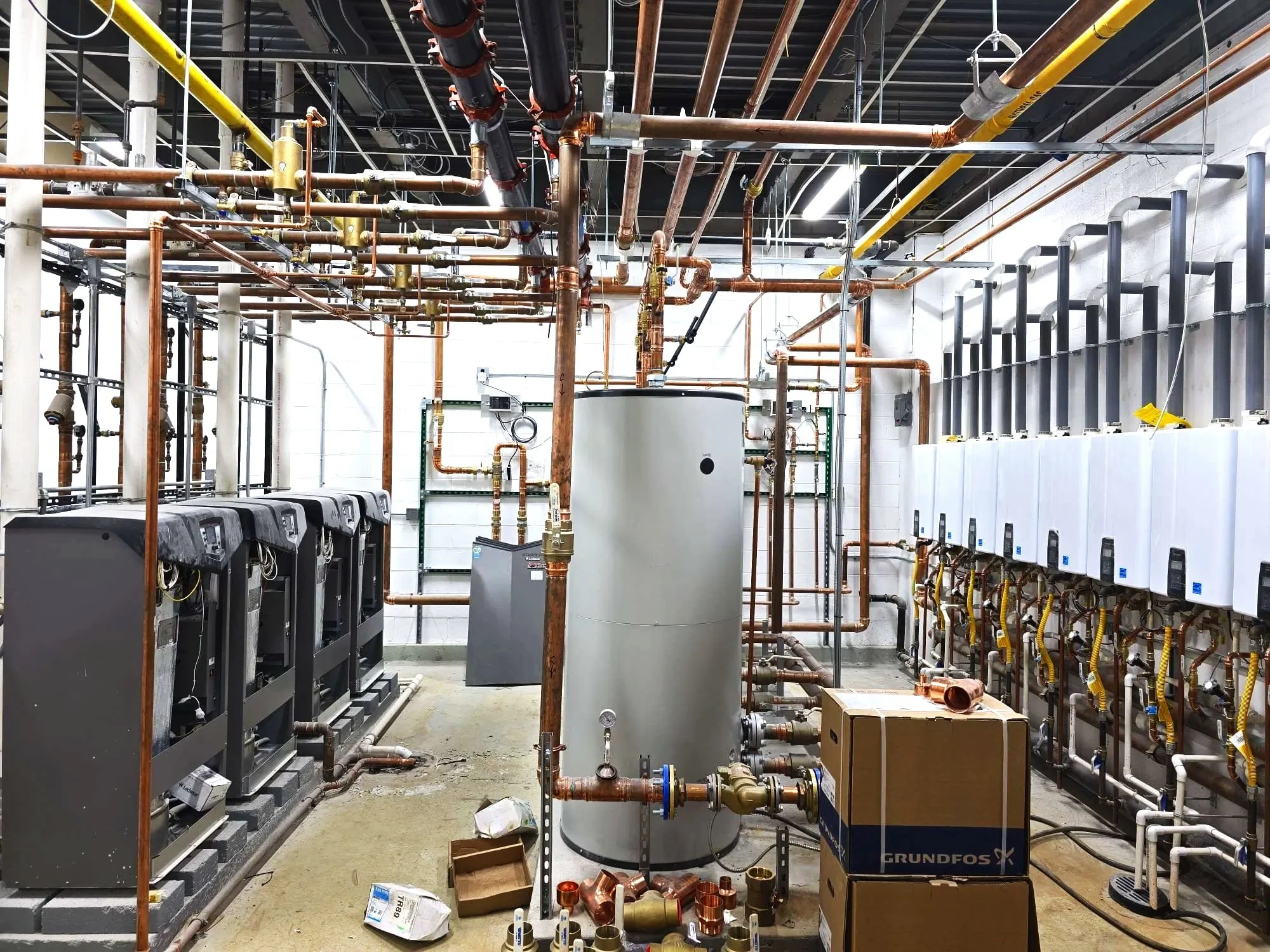 An electric water heater with pipes and valves, installed against a concrete wall.