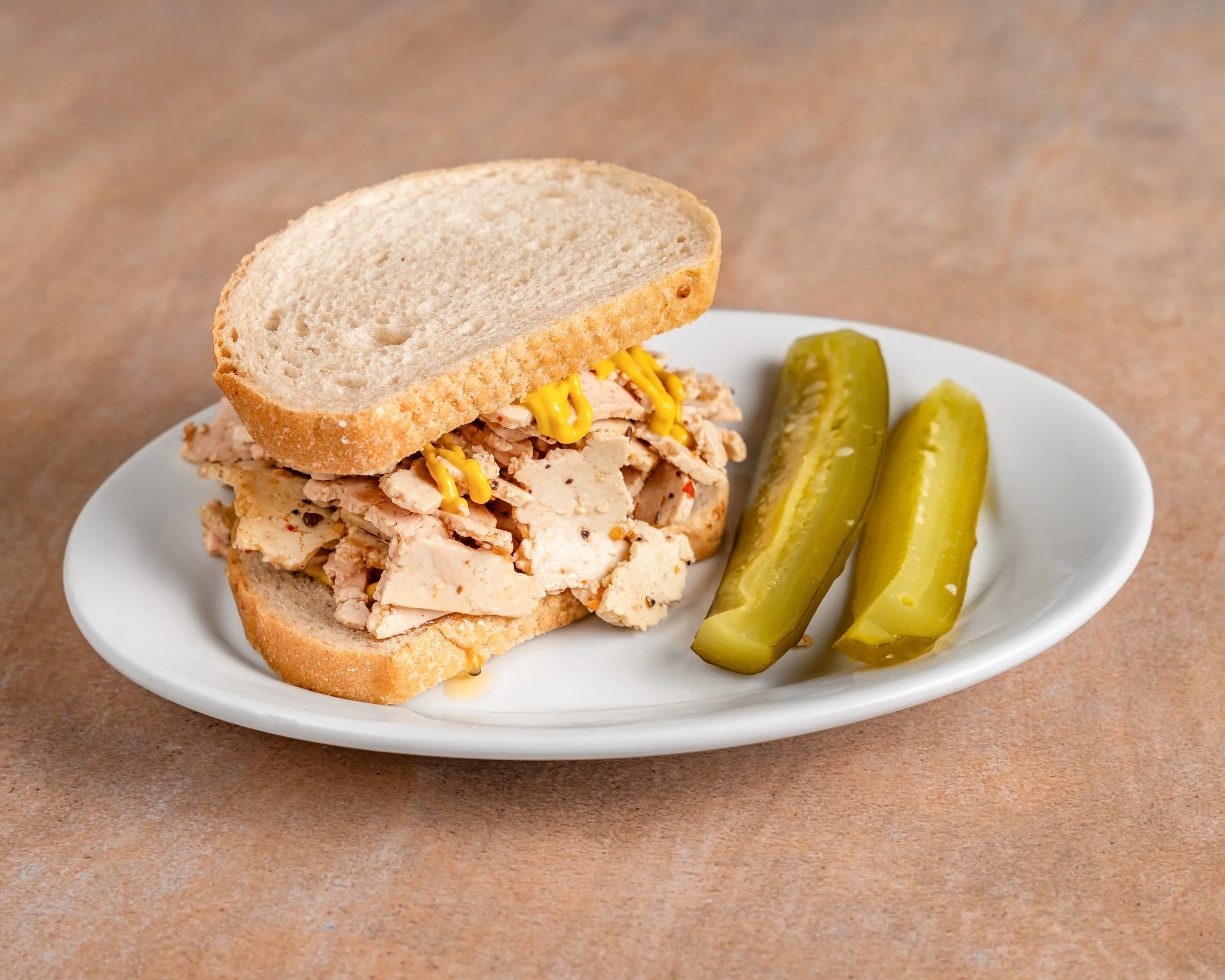 A plate with a tofu smoked meatsandwich with mustard and two pickle spears.