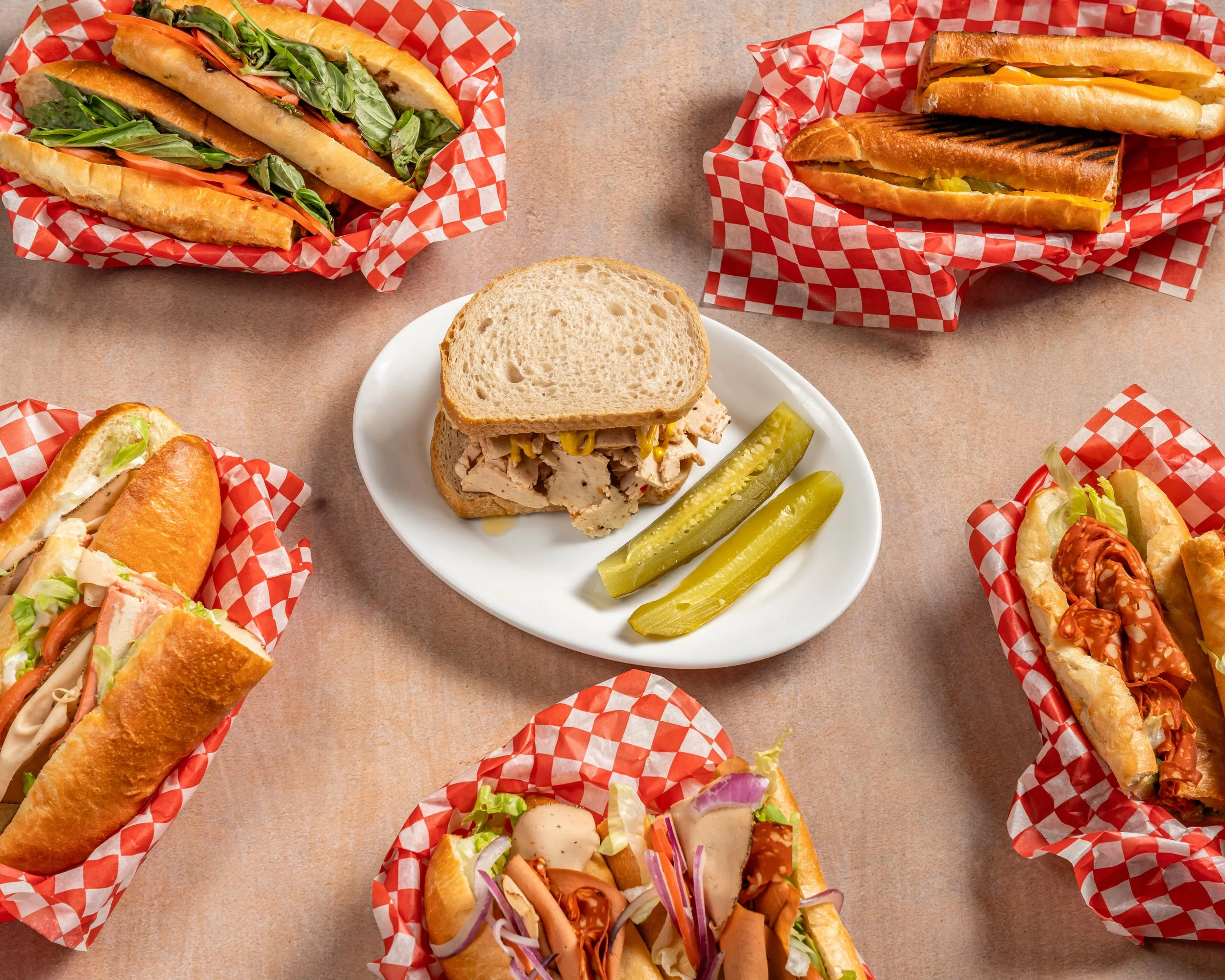 Several Vegan sandwiches along with pickle slices on a plate are arranged on a table with red and white checkered paper wrappers.