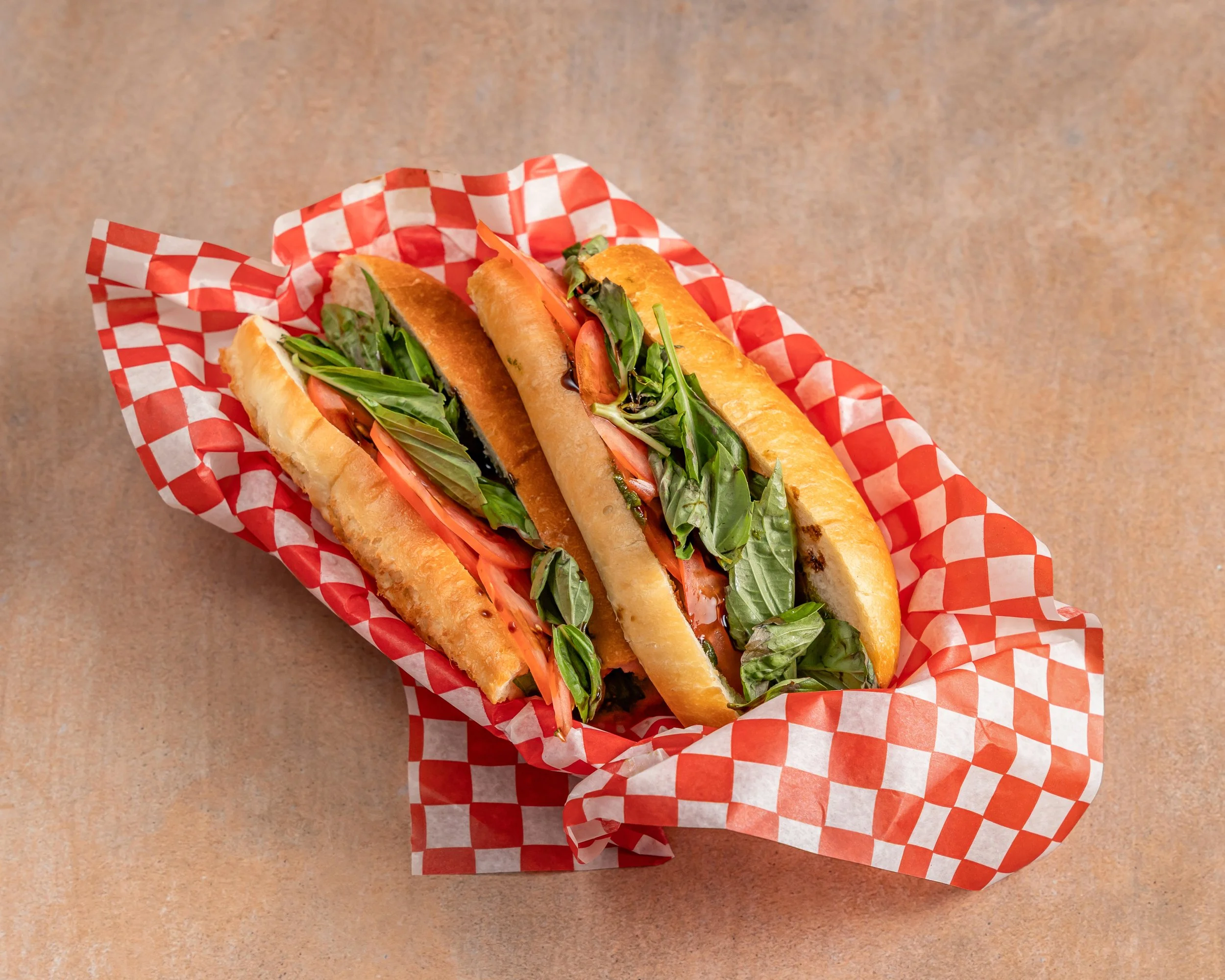 Two Italian hoagie sandwiches with fresh basil, tomato, and lettuce on a checkered red and white paper