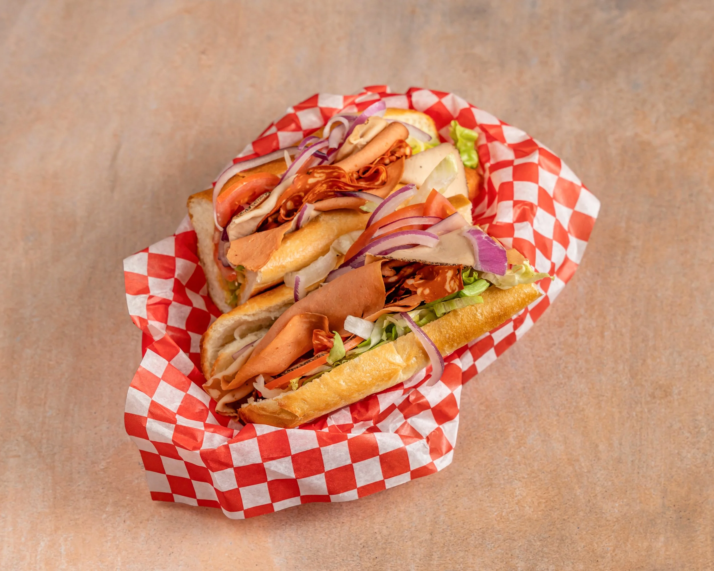 Two long sandwiches on a red and white checkered paper with sliced turkey, ham, lettuce, tomato, onions, and mayonnaise in a baguette style bread on a beige surface.