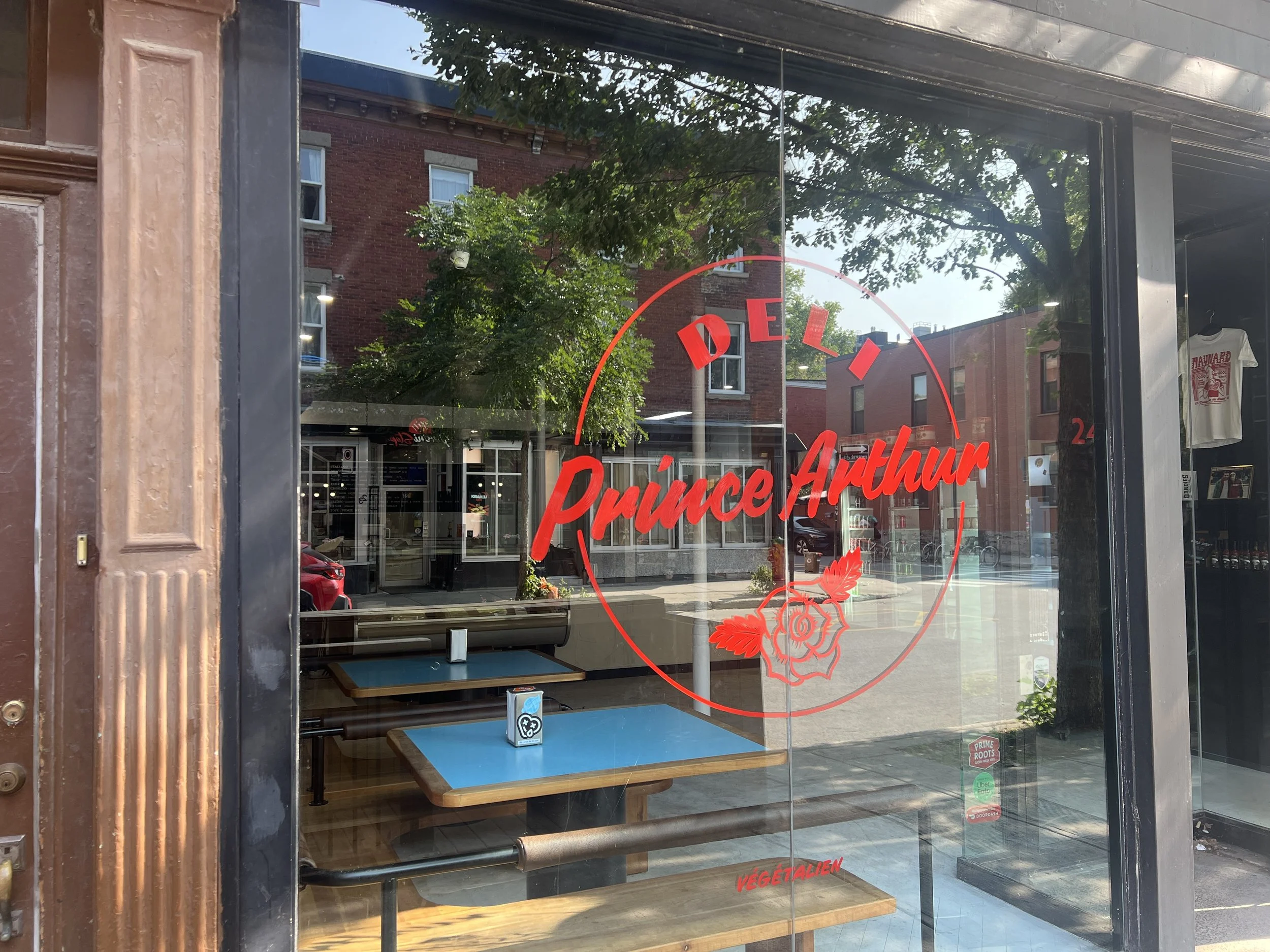 Restaurant window with red text reading "Prince Arthur" and a rose graphic, reflecting the street outside with trees and buildings.