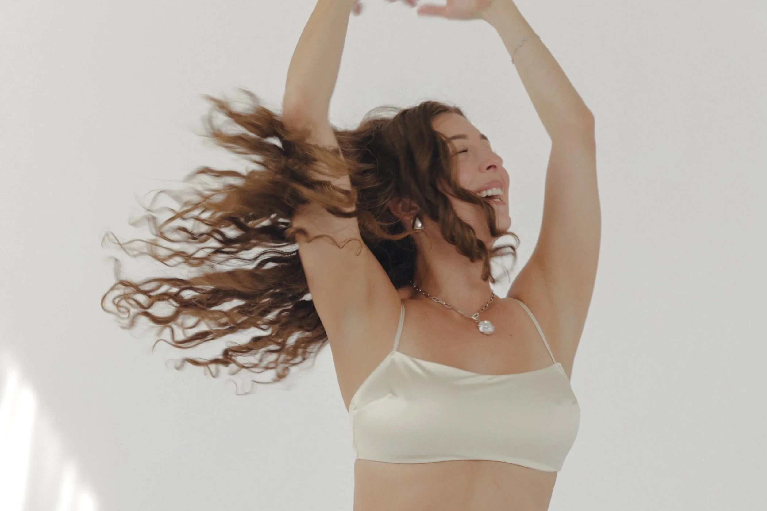 A woman with long curly hair dancing joyfully with her eyes closed, wearing a white satin camisole and a pearl necklace against a plain white background.