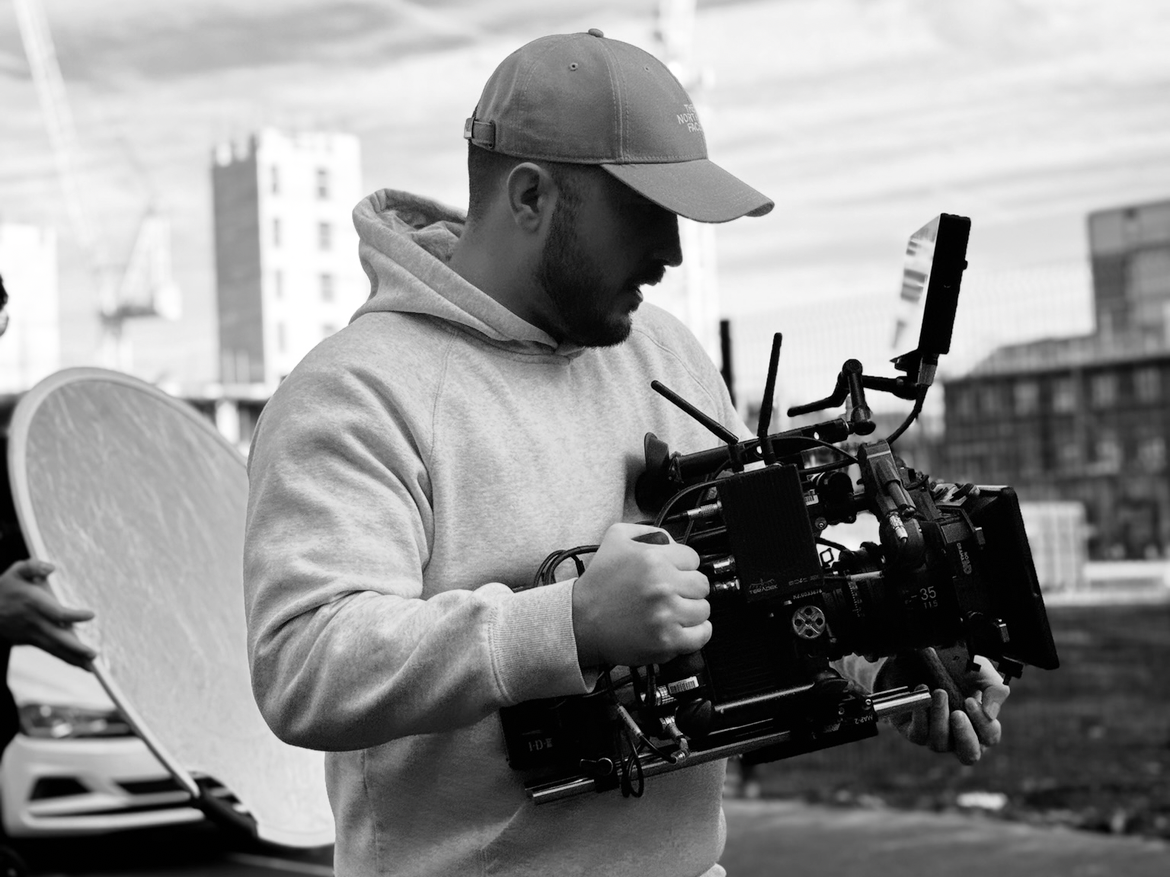 A man in a baseball cap and hoodie holding a professional camera rig on an outdoor set, with city buildings in the background.