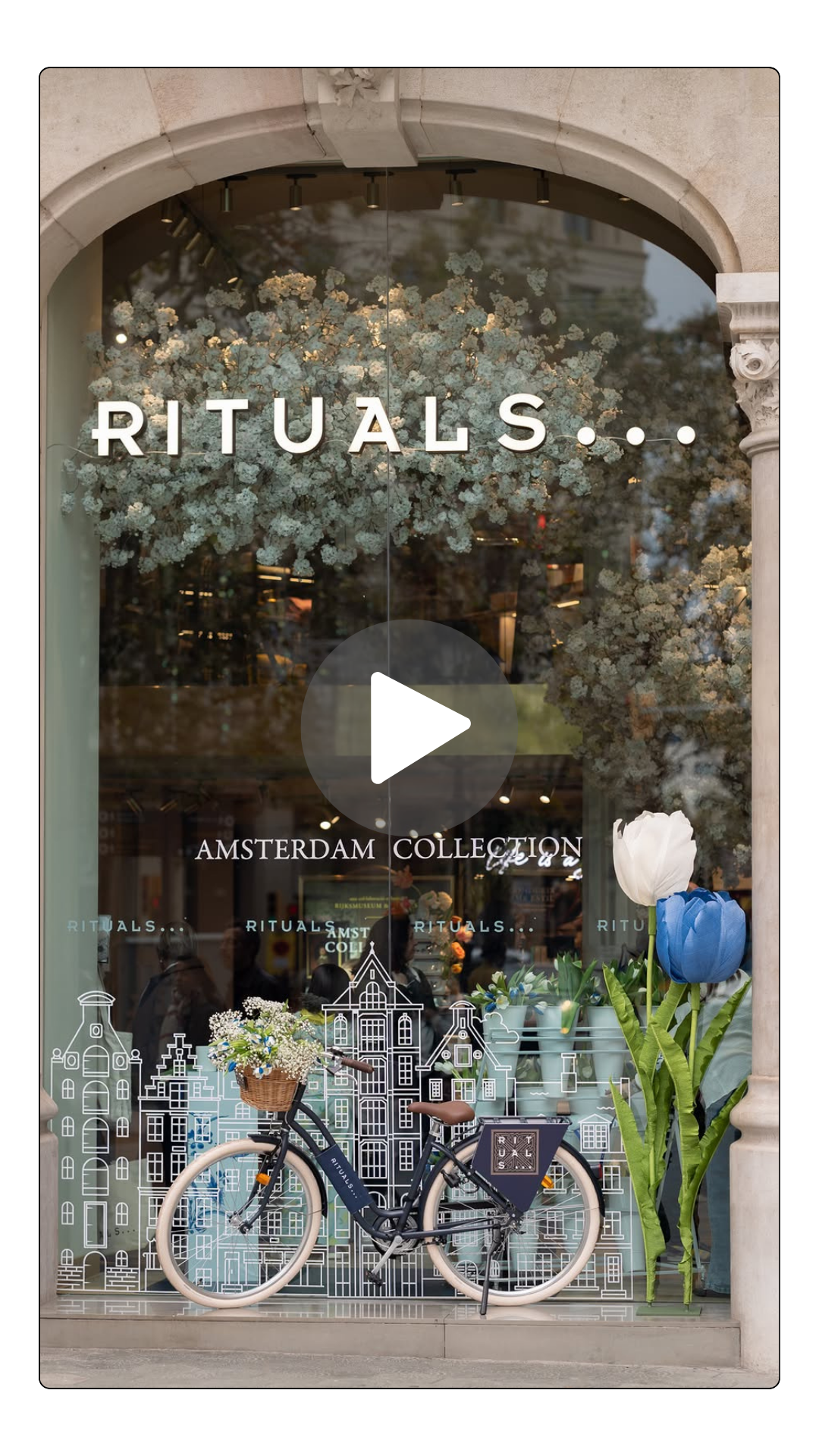 Storefront window with the sign 'RITUALS...' and 'AMSTERDAM COLLECTION', decorated with flowers, a bicycle with a basket of flowers, and large artificial tulips outside.