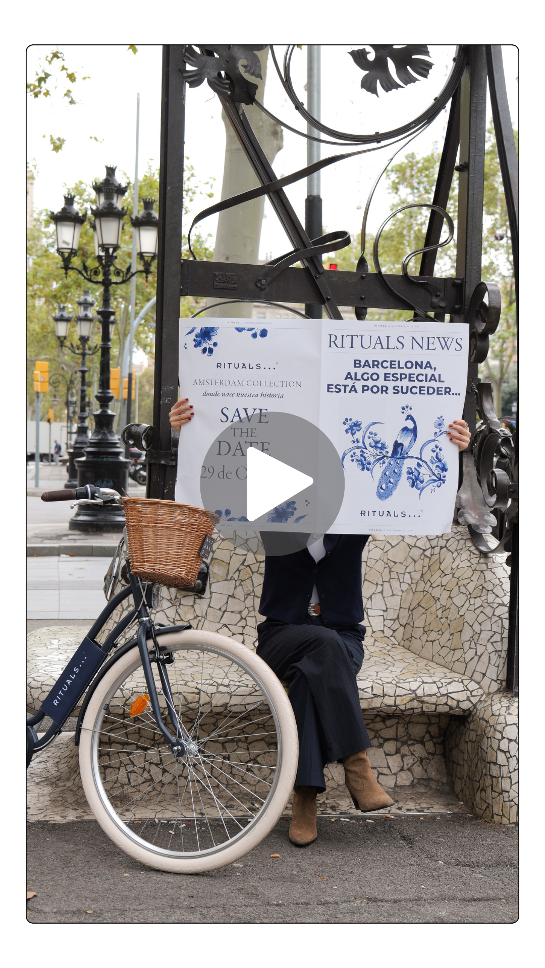 Person sitting on a mosaic bench holding a large sign with blue floral and peacock designs, and a bicycle with a front basket parked beside them on a city sidewalk.