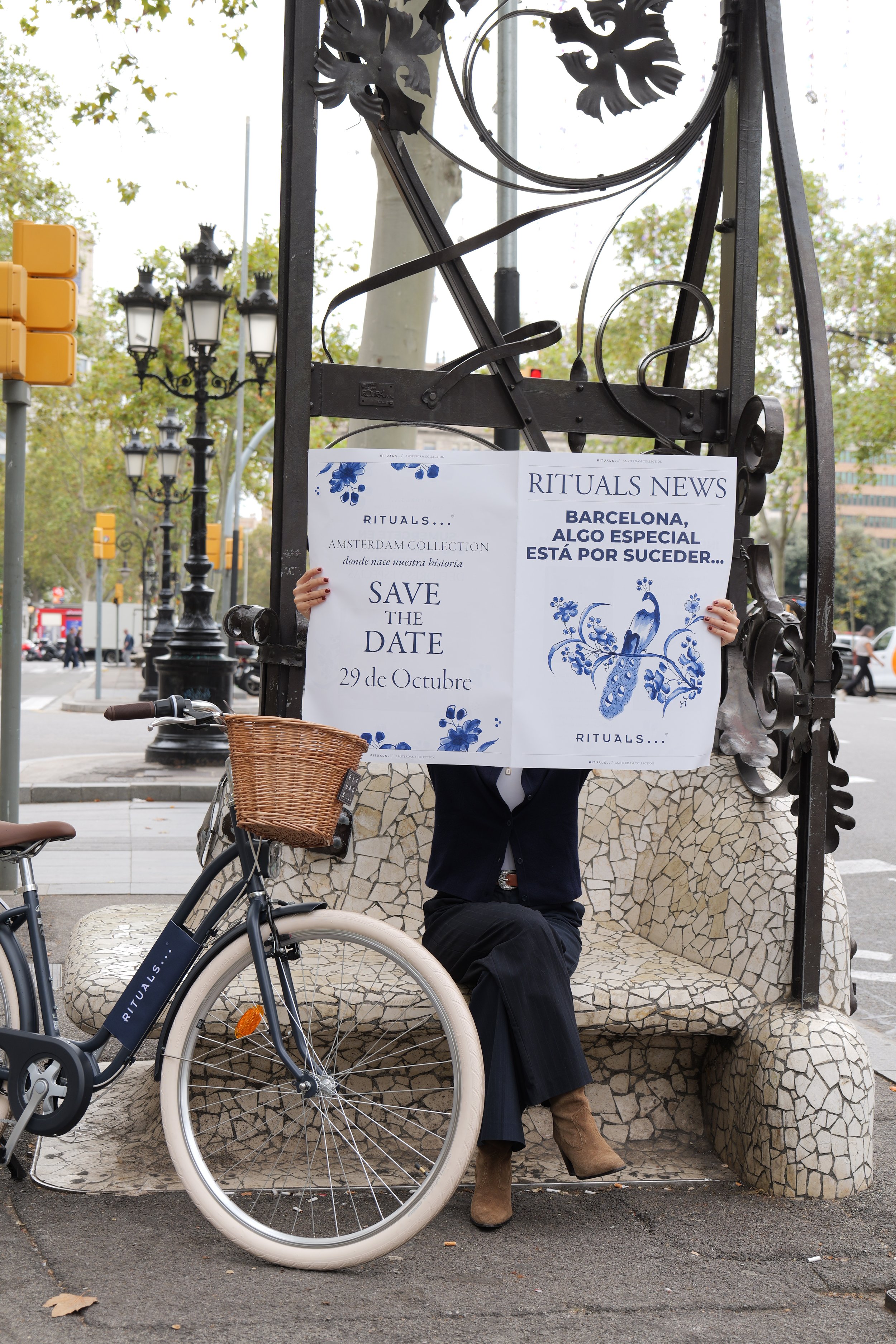 Person sitting on mosaic bench holding signs advertising upcoming event in Barcelona, with bicycle parked nearby.